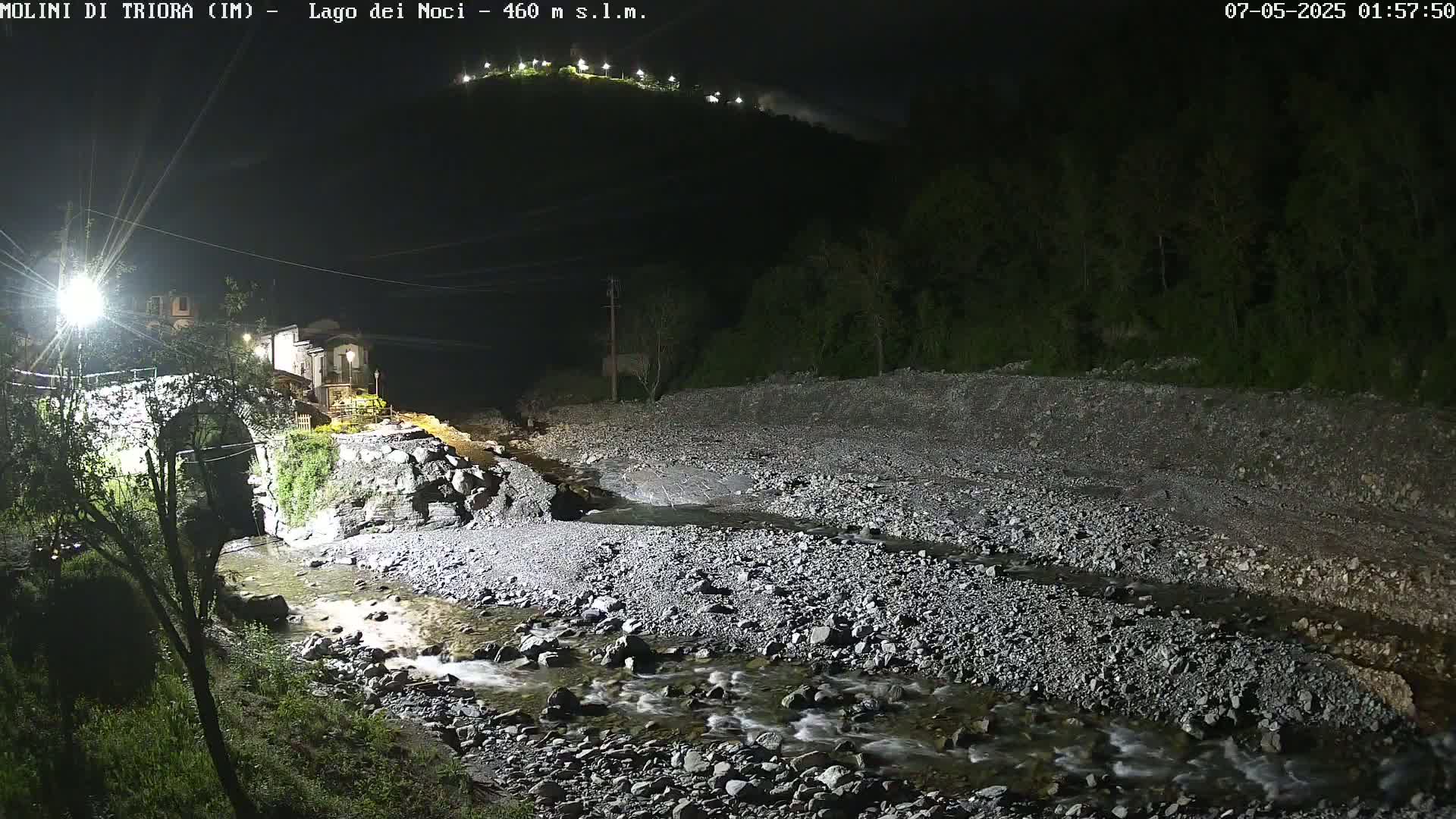 A rocky, shallow stream flows at night past a stone bridge and buildings, under a dark sky illuminated by lights on a distant hill.