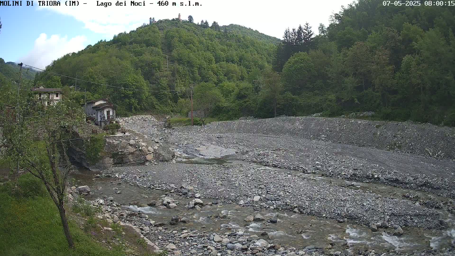 A rocky riverbed with a small stream flows through it, nestled between green hills under a partly cloudy sky.