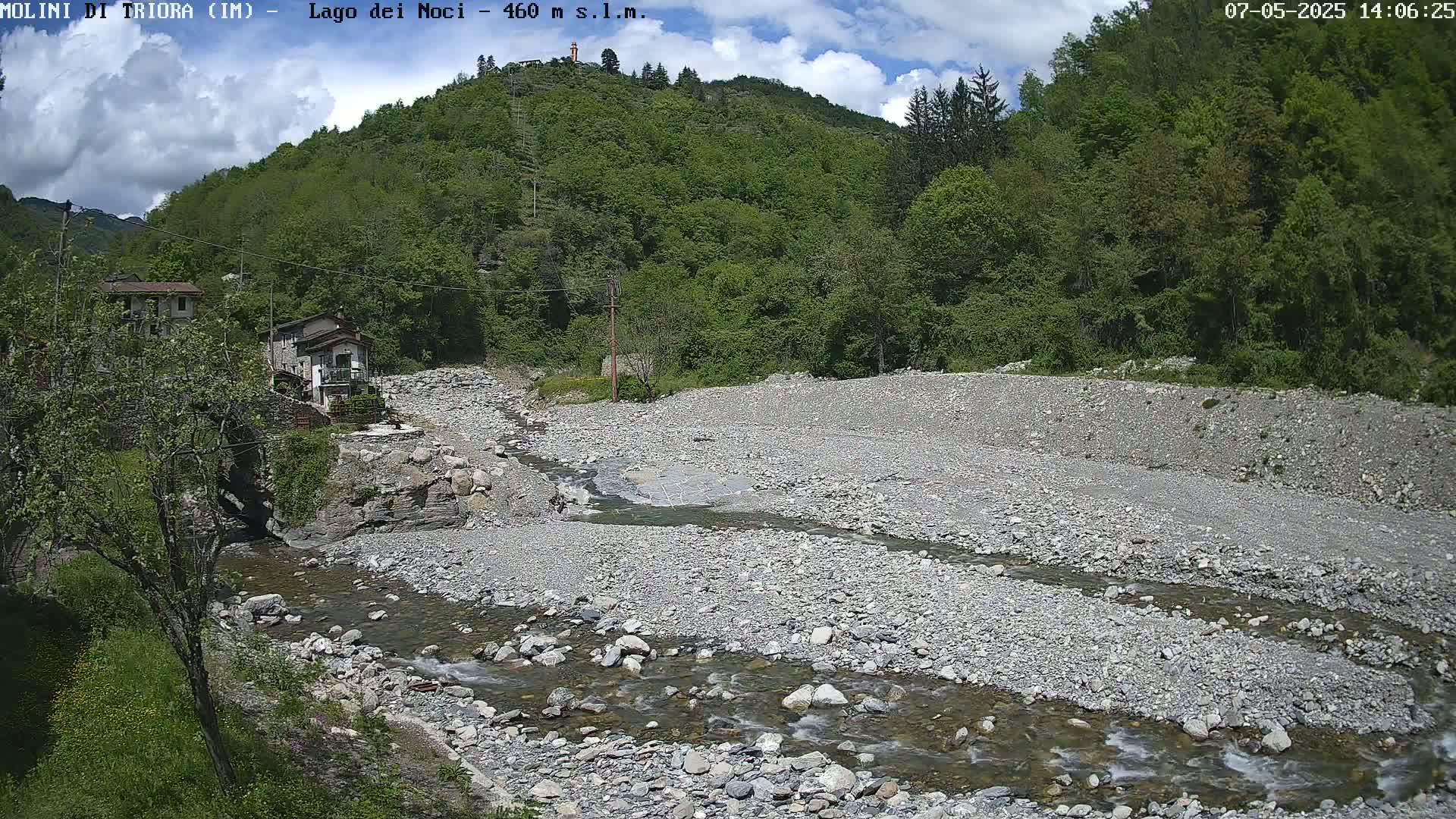 A rocky riverbed with a shallow stream flows through it, past some buildings nestled against a lush green hillside under a partly cloudy sky.