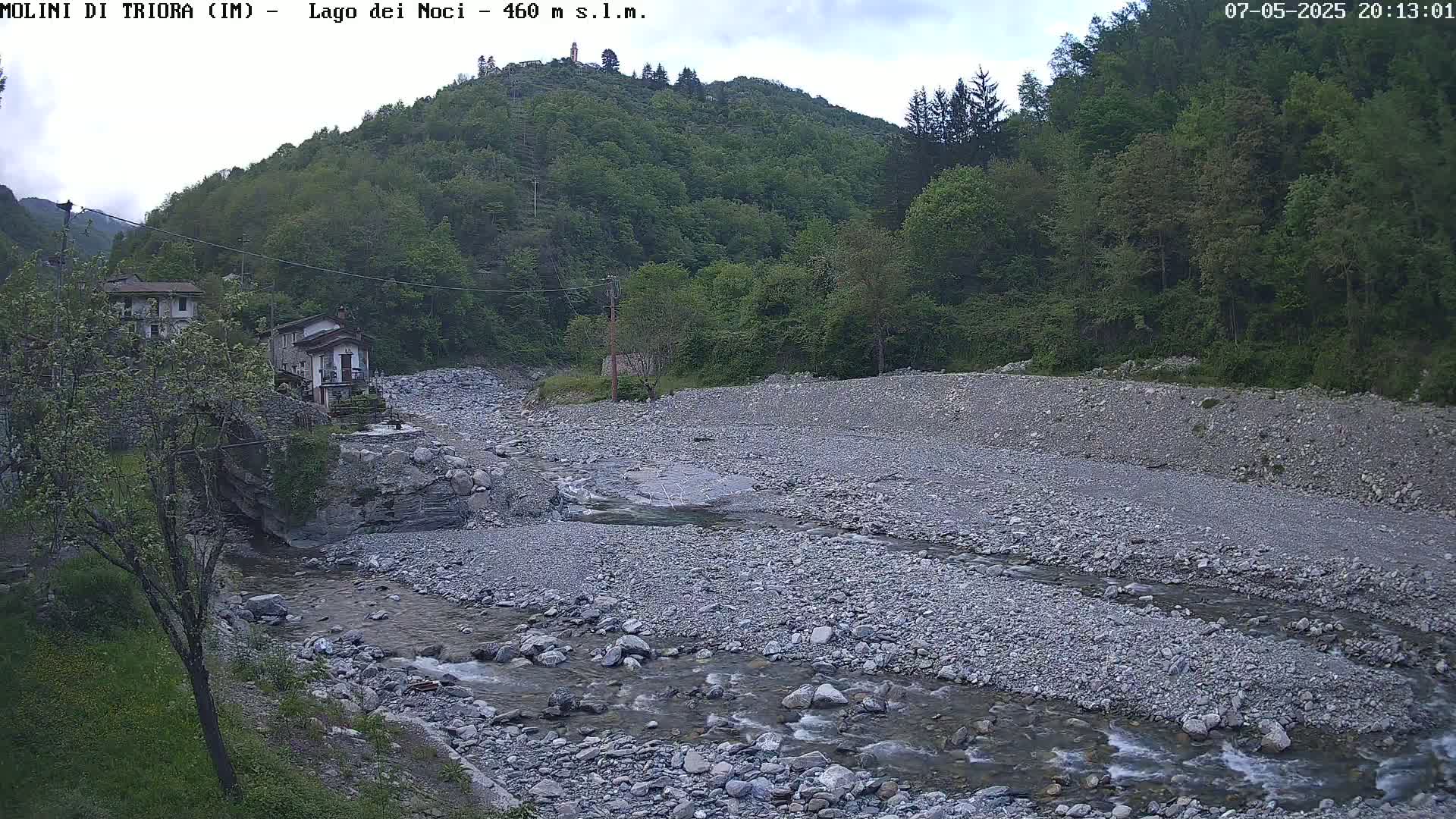A shallow, rocky river flows past two buildings nestled in a green, hilly landscape under a partly cloudy sky.