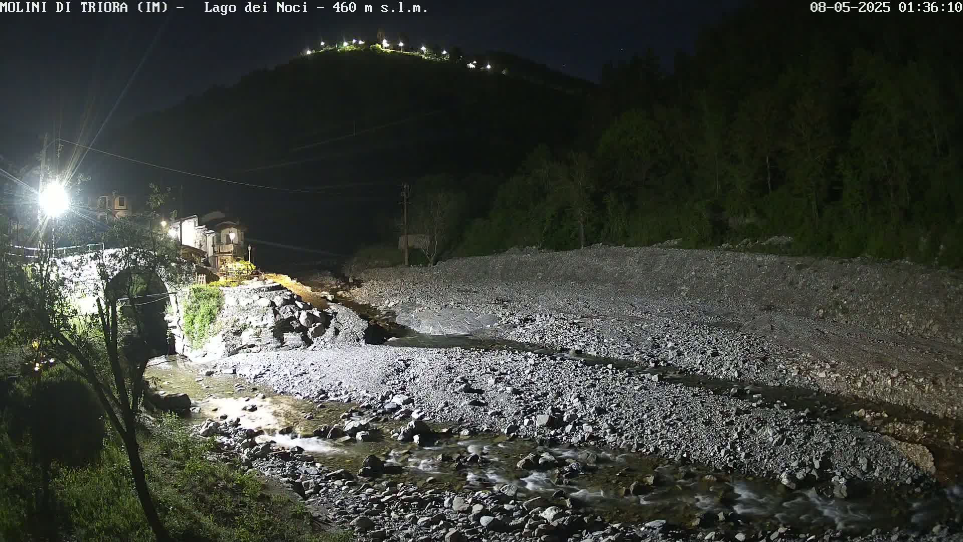 A rocky stream flows under a stone bridge at night, past a small village nestled at the foot of a dark, hilltop.