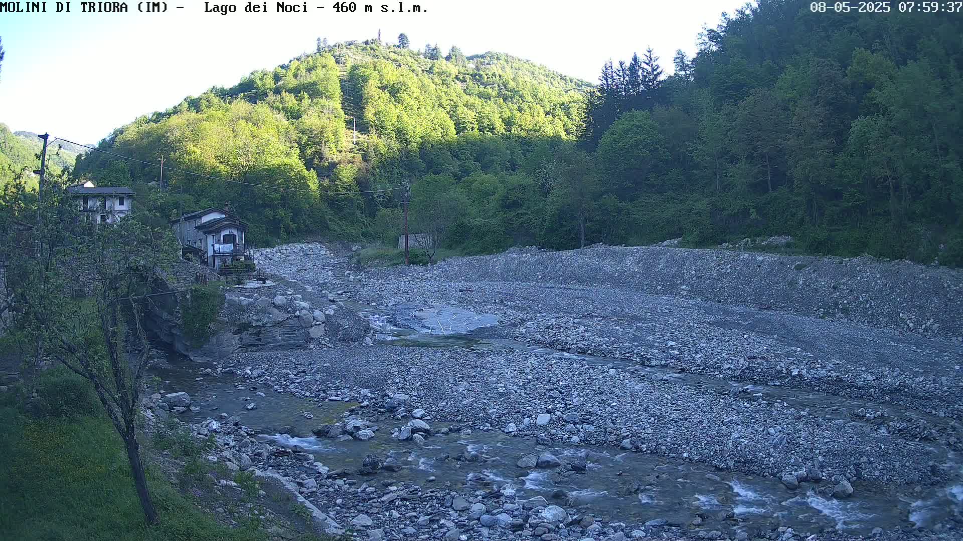 A rocky riverbed with a shallow stream flows through a valley under a clear sky, past two buildings nestled among lush green hillsides.