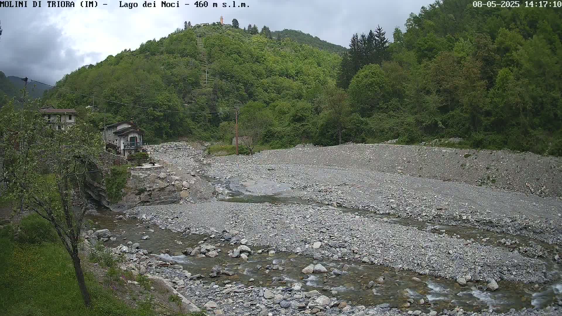 A rocky stream flows through a dry riverbed, past buildings nestled amongst lush green hills under a cloudy sky.
