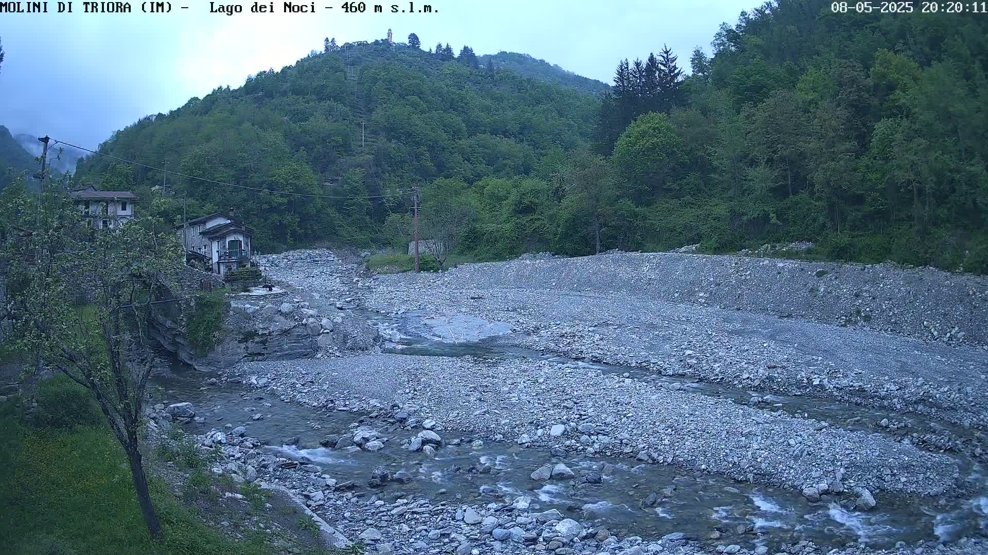 A rocky river flows through a valley under an overcast sky, past some buildings nestled amongst lush green hills.