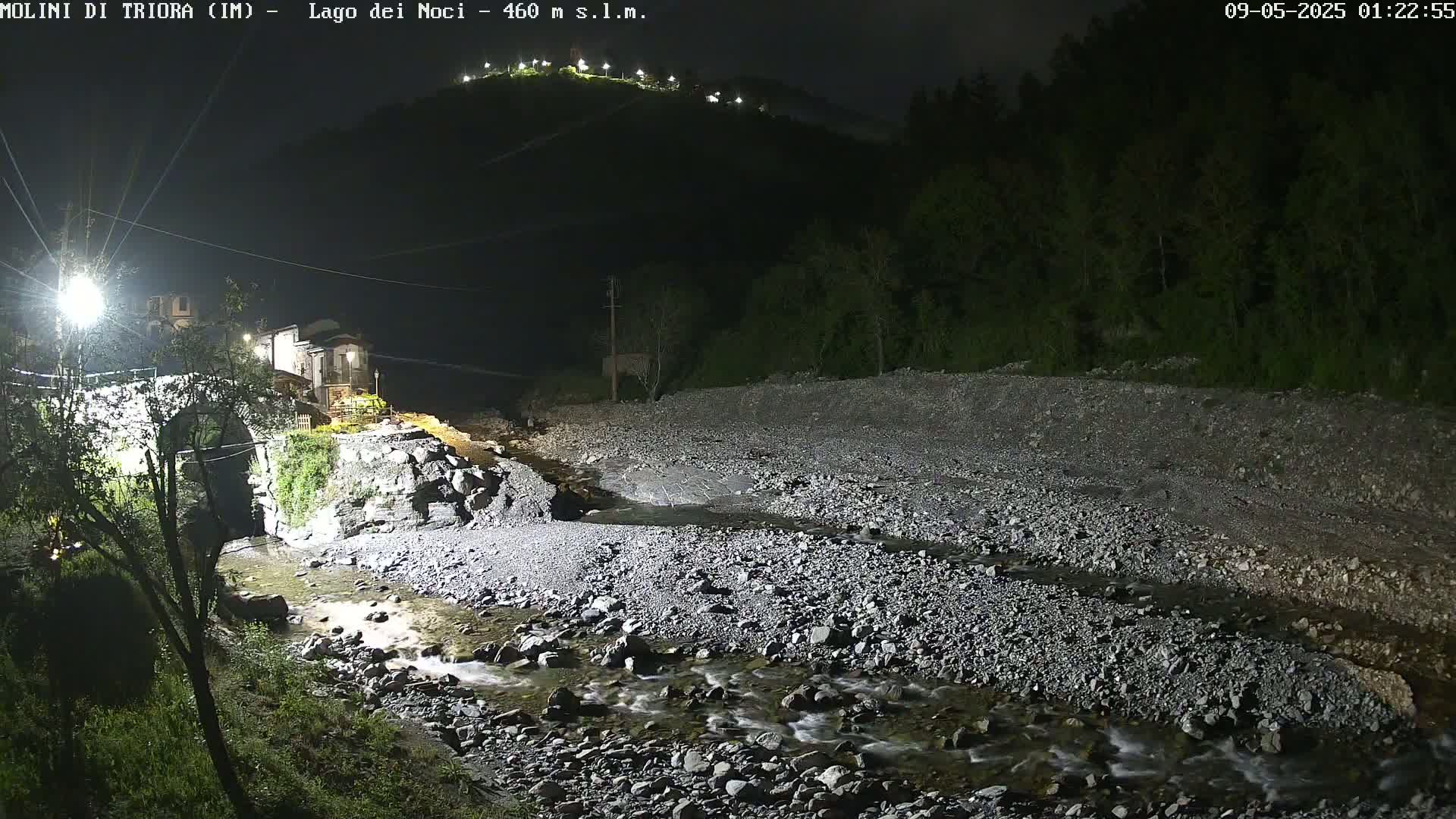 A rocky stream flows under a stone bridge at night, past a small village nestled against a dark hillside dotted with lights.