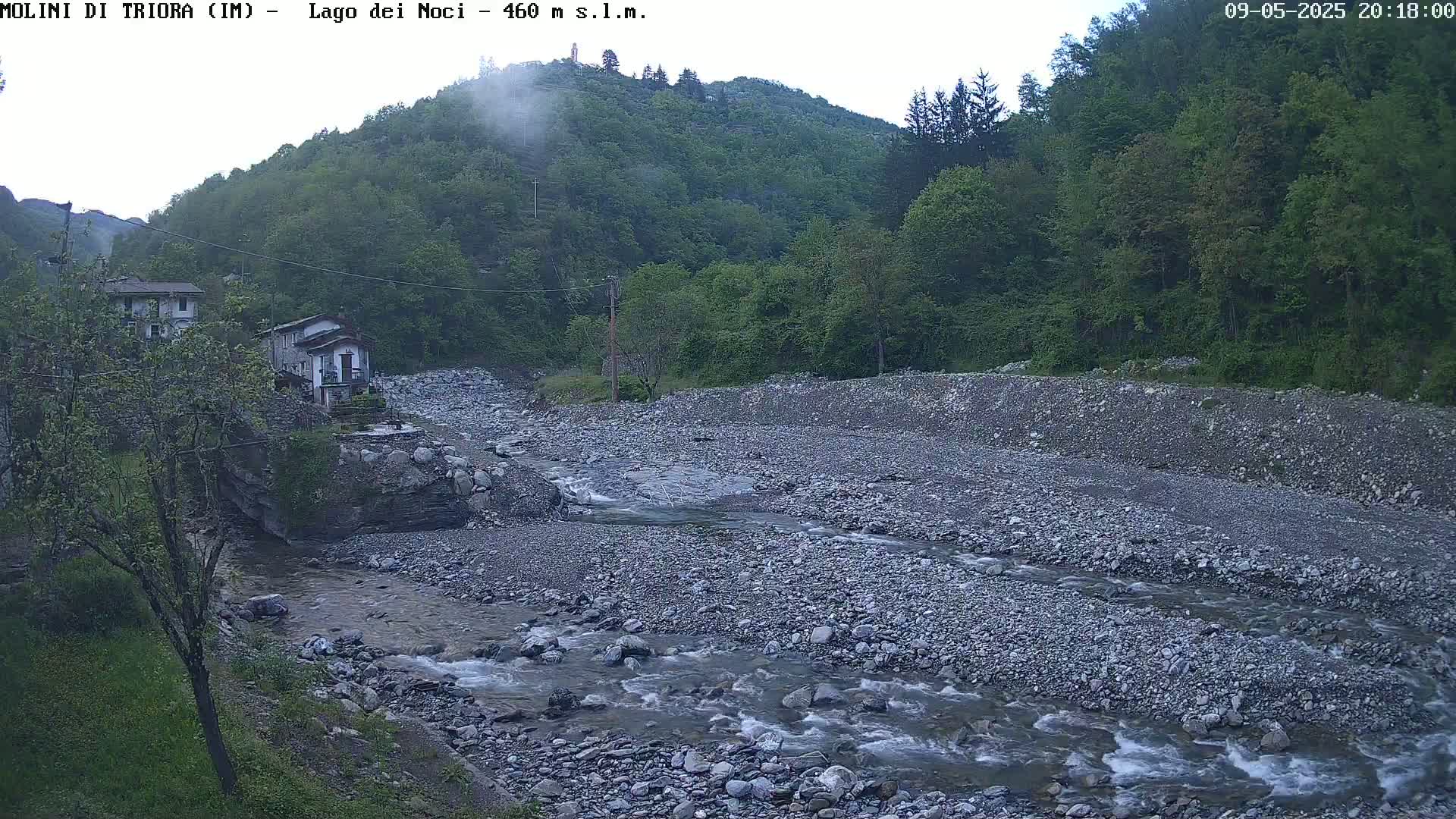 A rocky river flows through a valley, alongside a few buildings and lush green hills under an overcast sky.