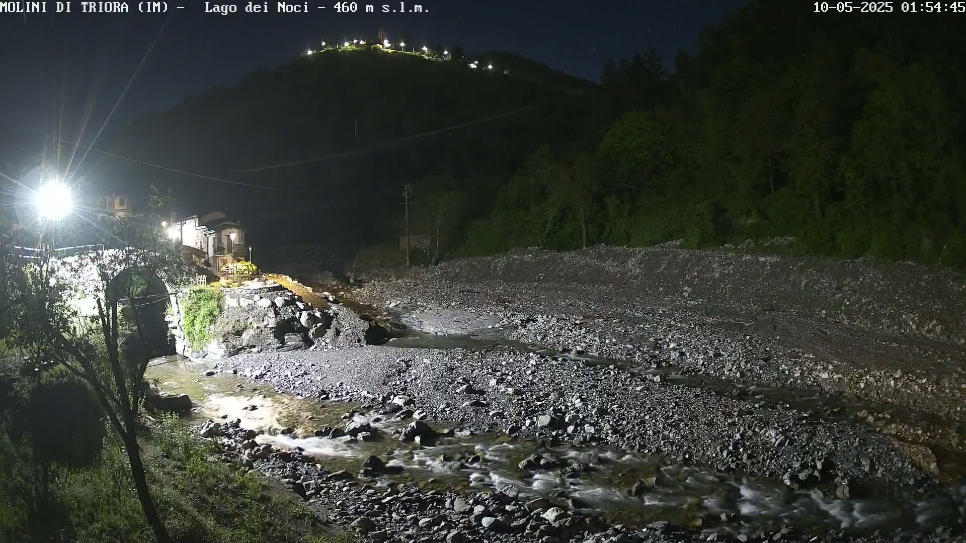 A rocky stream flows under a bridge near a village at night, under a clear sky.