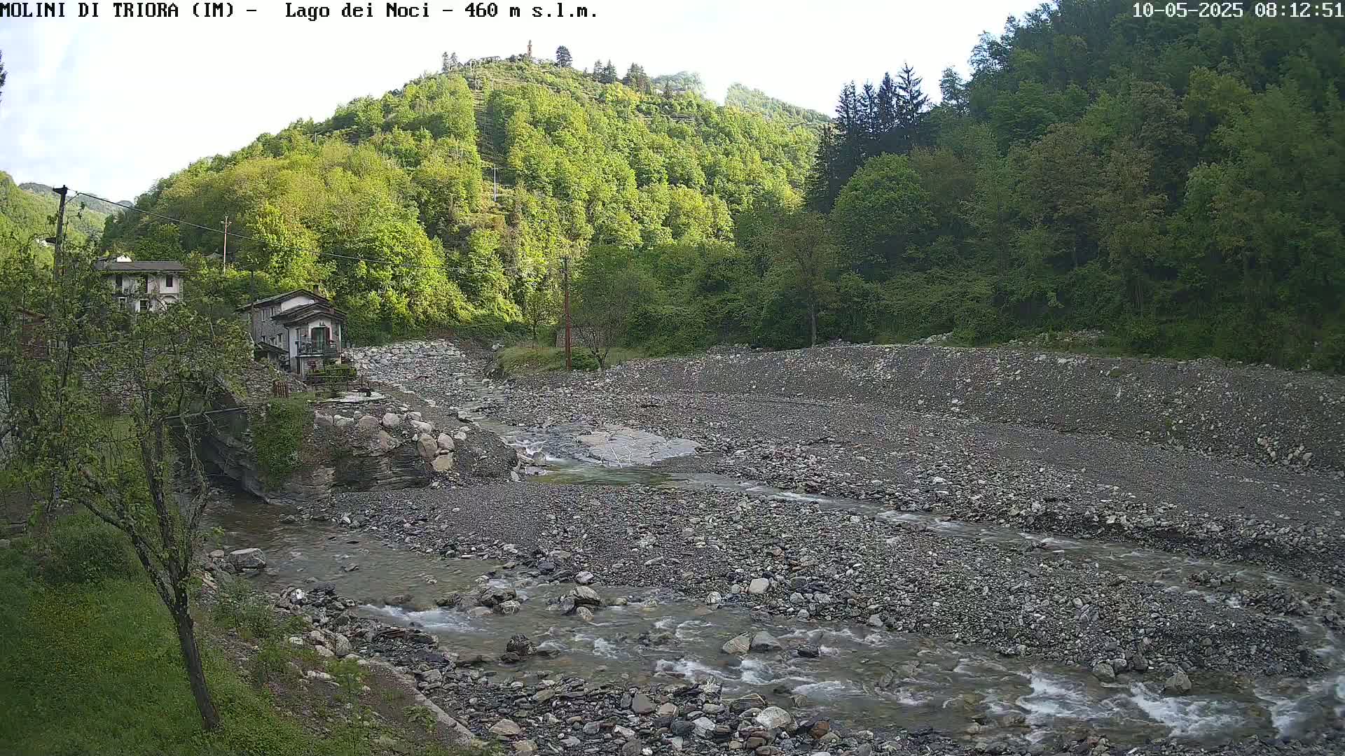 A rocky river flows through a dry riverbed, past a couple of buildings nestled in a lush green hillside under a mostly clear sky.