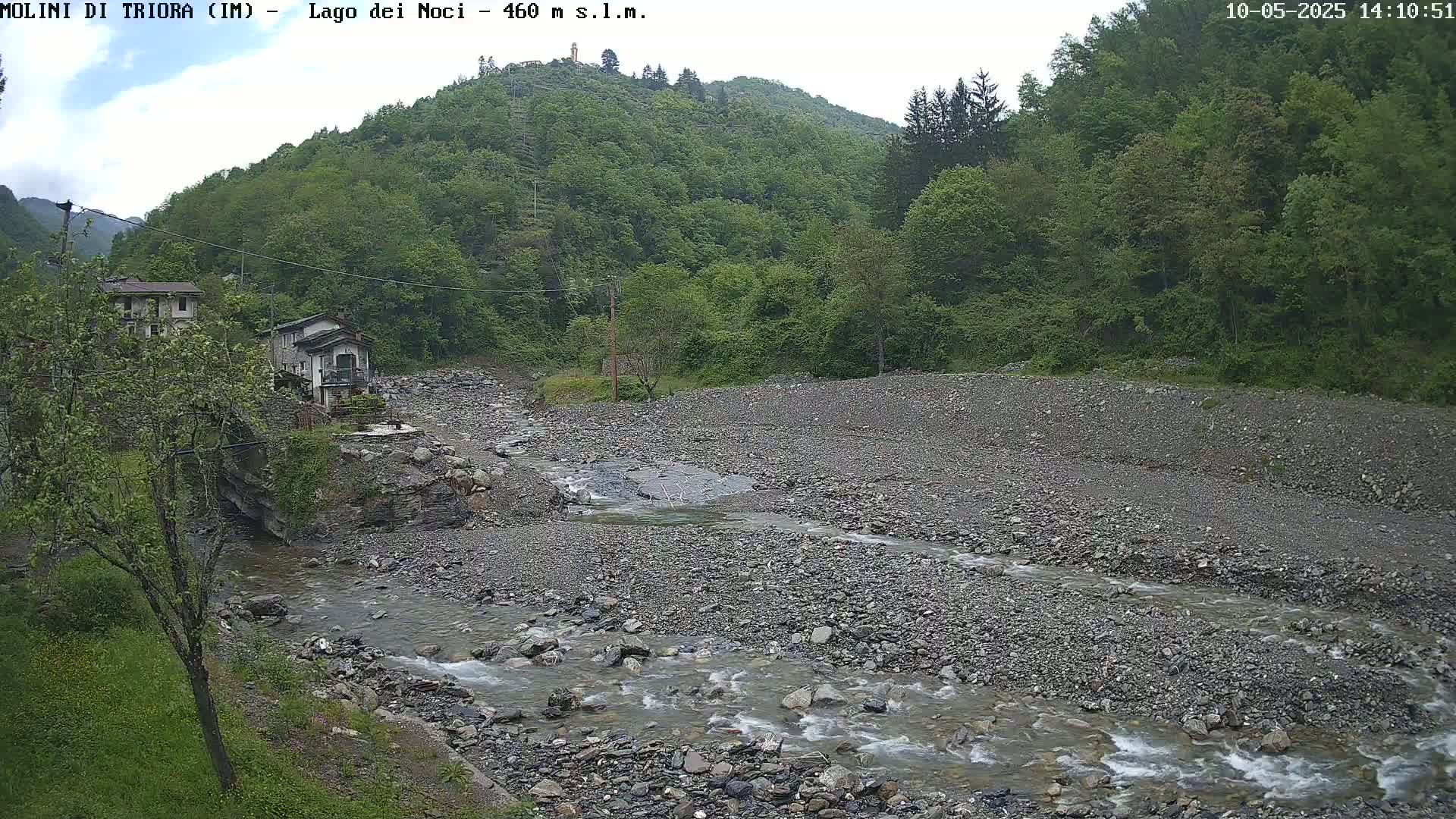 A rocky stream flows through a gray, pebbly riverbed, past some buildings nestled beside a lush green hillside under an overcast sky.