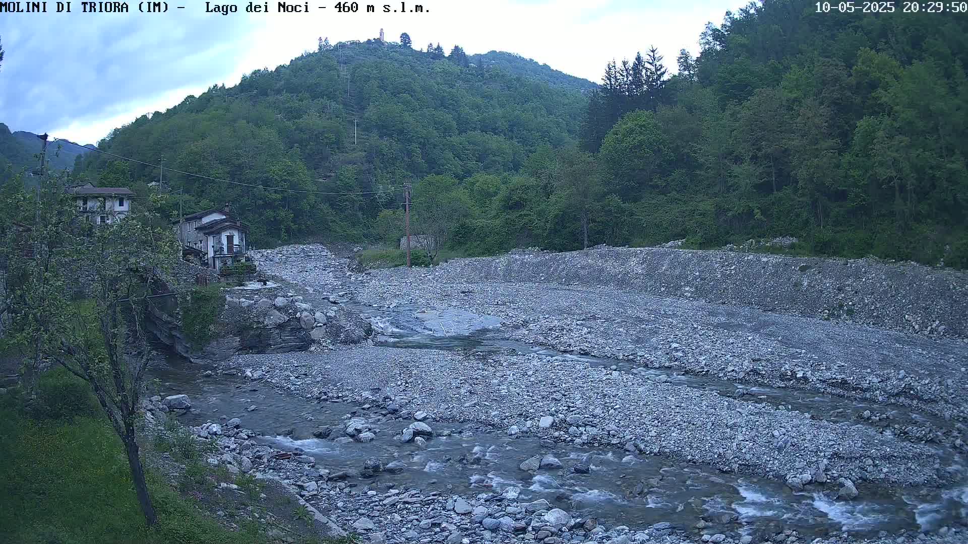 A rocky river flows through a valley under an overcast sky, past a small village nestled against a lush green hillside.