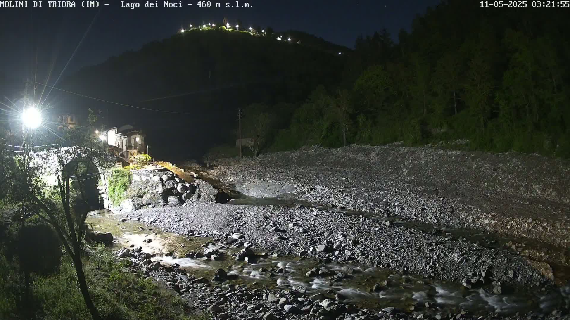 A rocky, shallow river flows under a stone bridge at night, past a small village nestled at the base of a dark, tree-covered hill illuminated by lights.