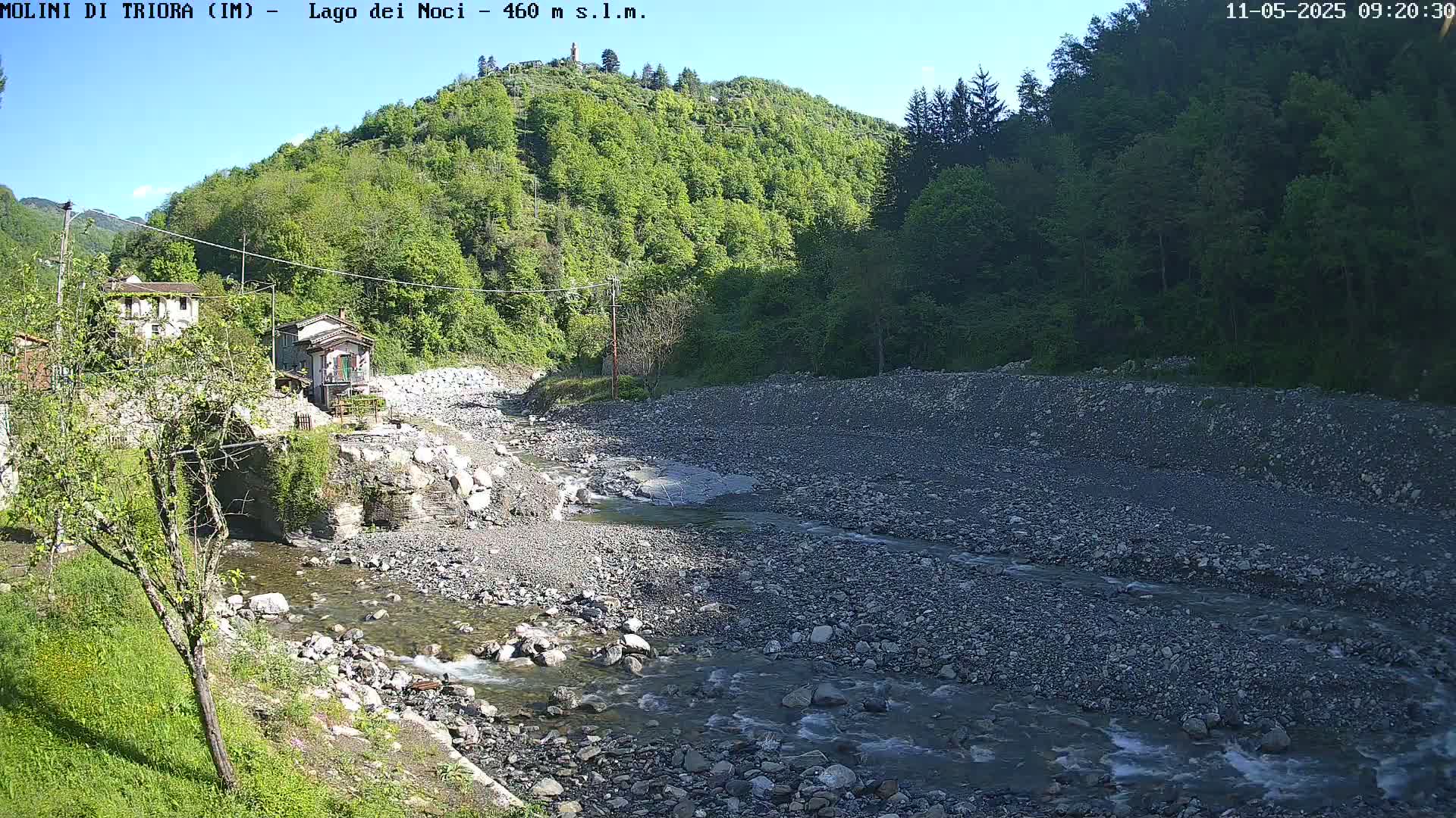A rocky river flows through a valley under a clear blue sky, past a stone bridge and buildings nestled among lush green hills.