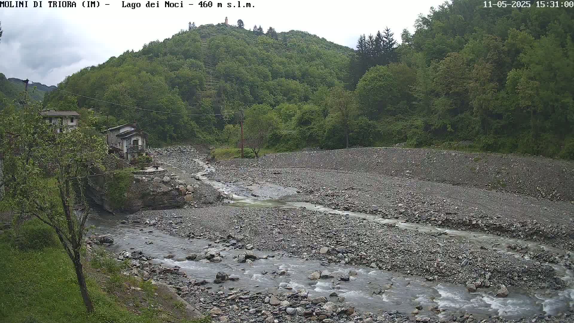 A rocky river flows through a valley, past a pair of buildings nestled among lush green hills under an overcast sky.