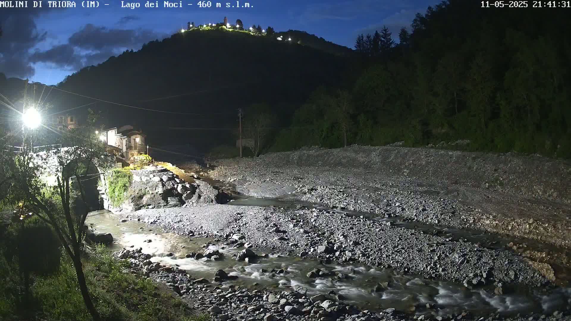 A rocky river flows under a stone bridge near a small village at night, under a partly cloudy sky.