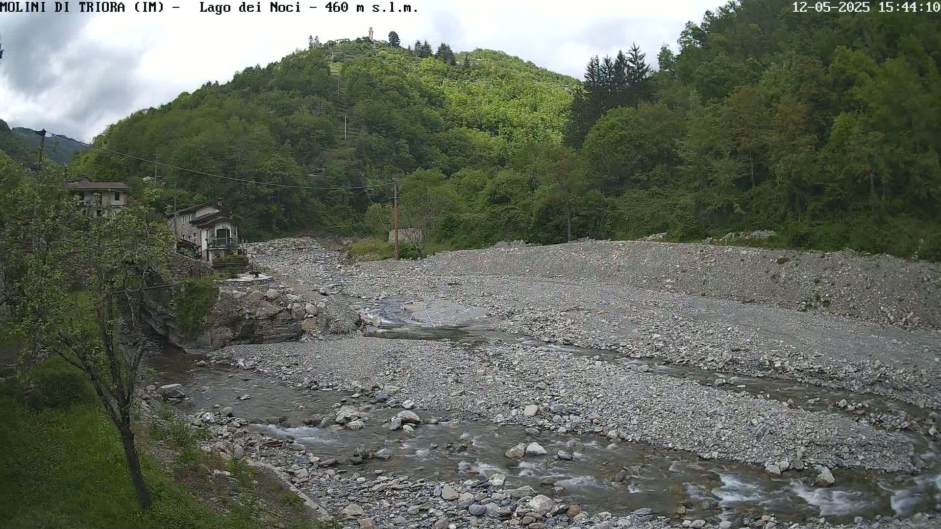 A rocky river flows through a valley under a partly cloudy sky, past a couple of buildings nestled in lush green hills.