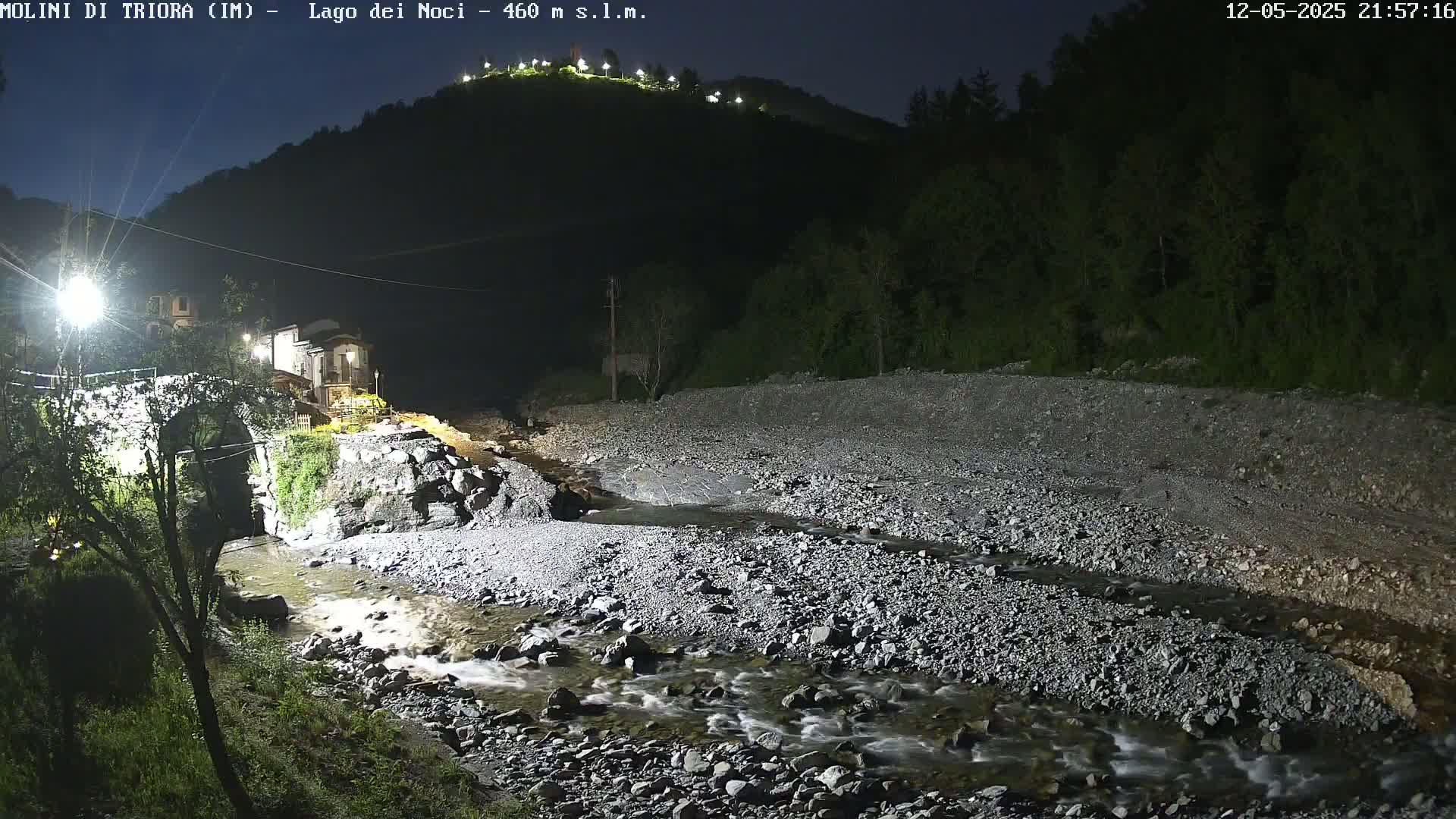 A rocky river flows through a dark, nighttime valley illuminated by artificial lights, past a stone bridge and village nestled against a hillside.