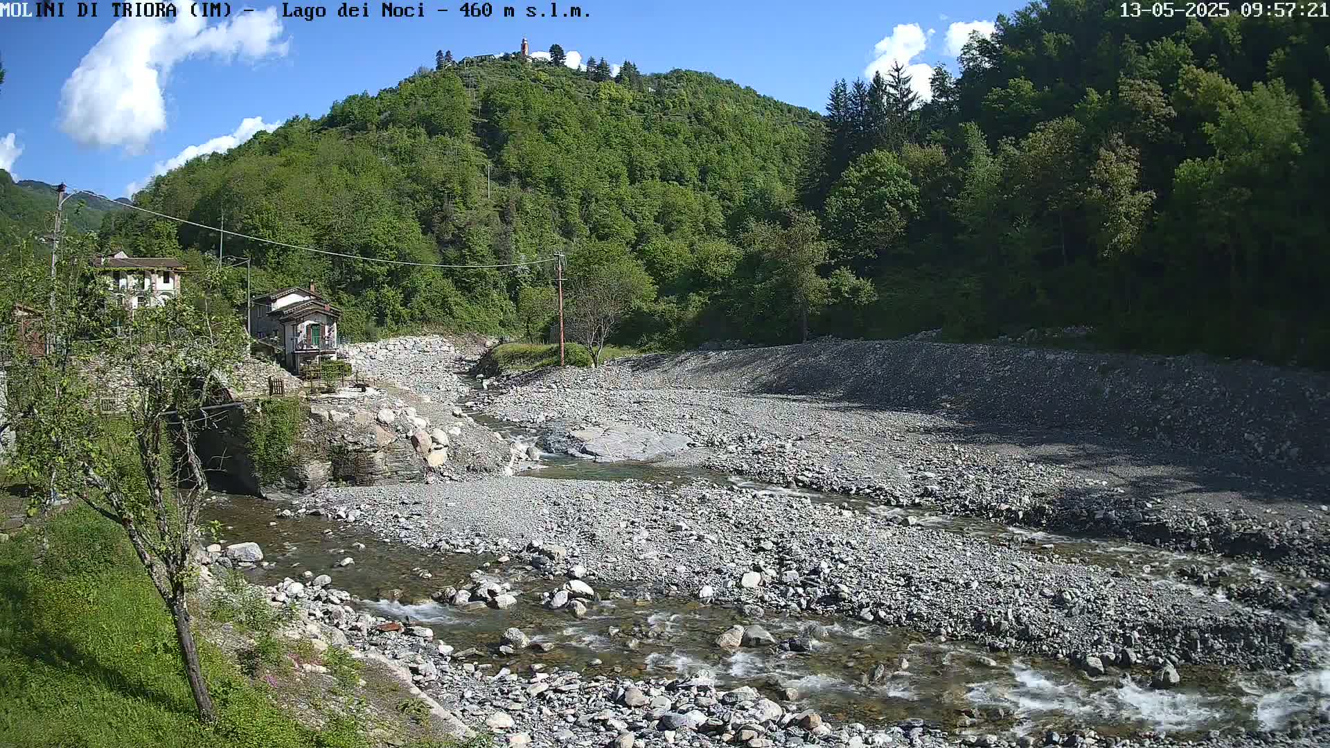 A rocky, shallow river flows through a valley under a partly cloudy sky, past stone buildings and lush green hills.