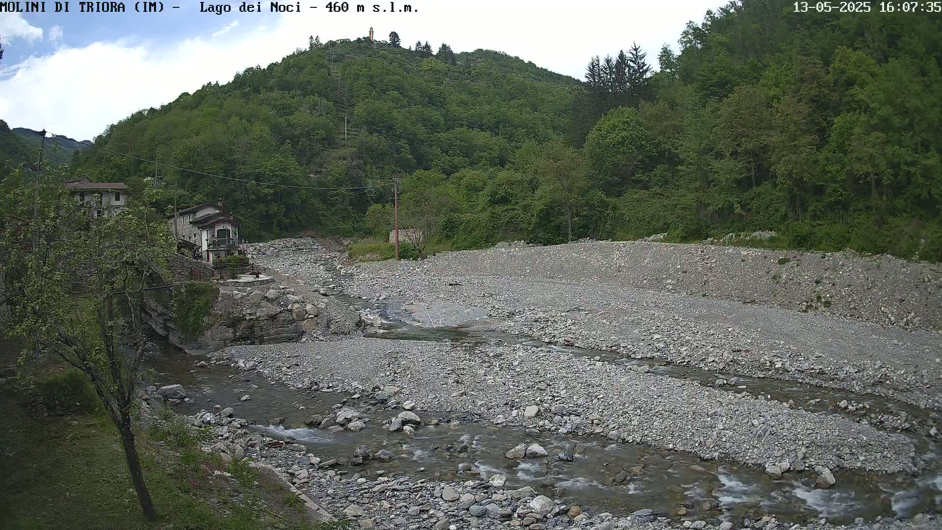 A rocky riverbed with a shallow stream flows through it, past a small village nestled against a lush, green hillside under a mostly cloudy sky.