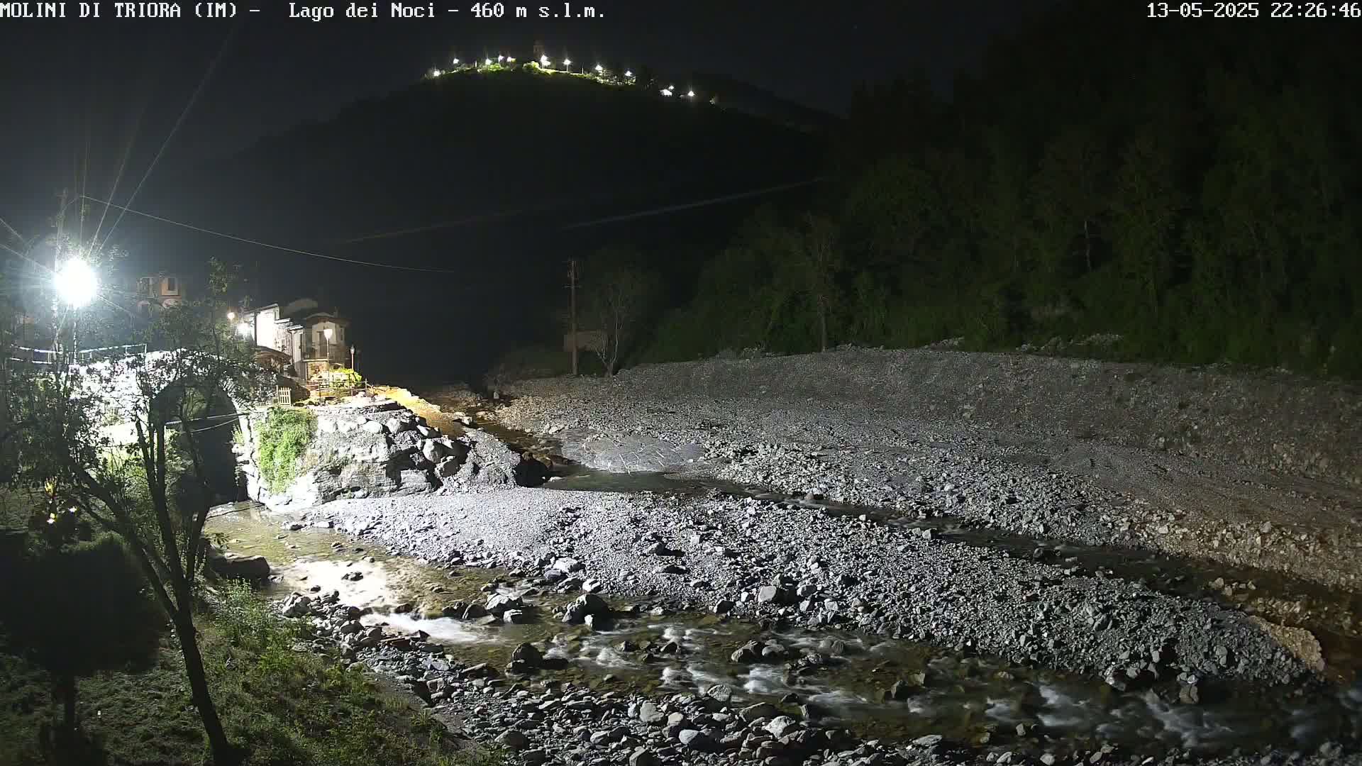 A rocky stream flows under a stone bridge at night, past buildings illuminated by artificial light, with a hilltop visible in the background under a dark sky.