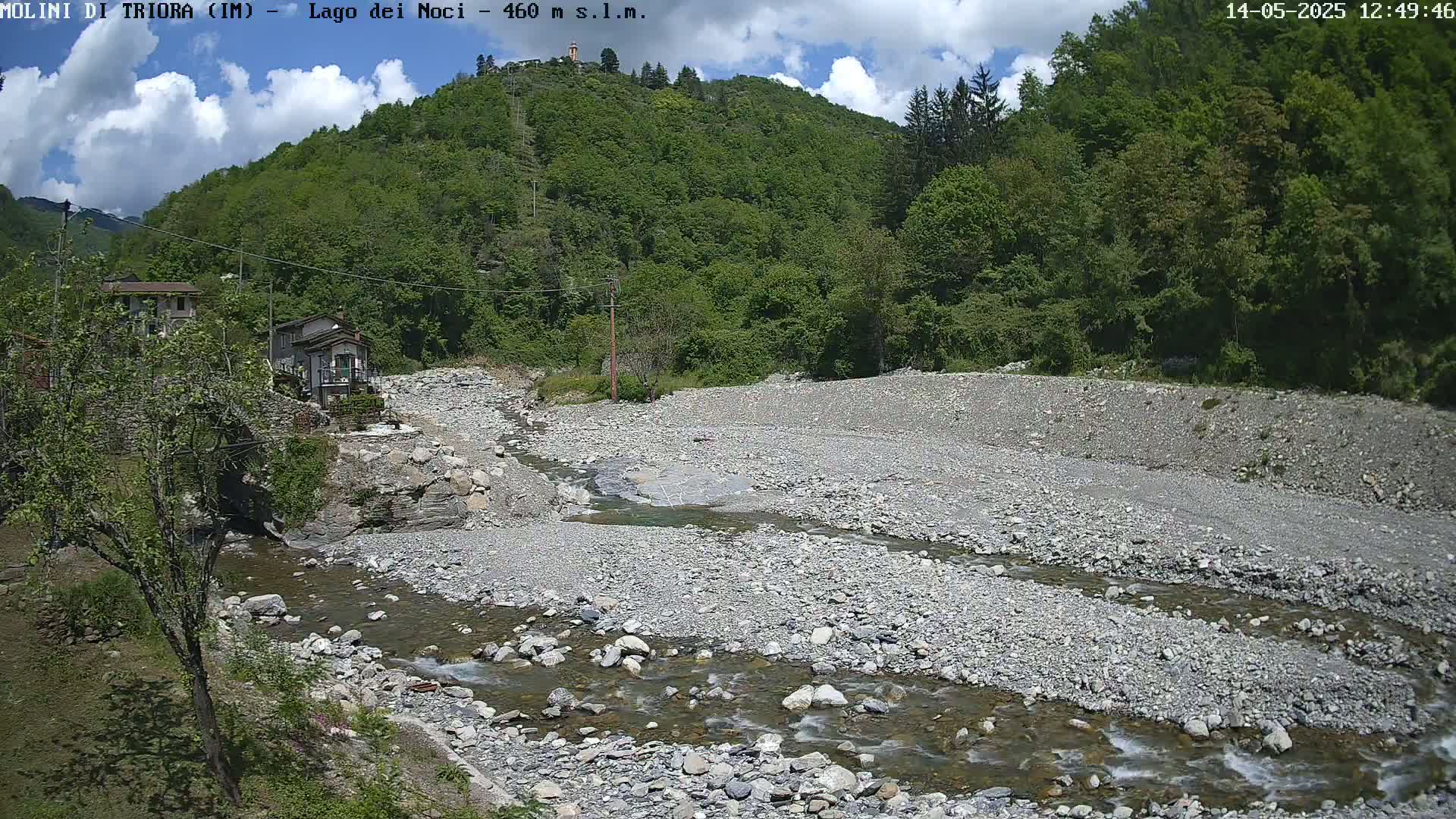 A rocky, shallow river flows through a dry riverbed, past a few buildings nestled at the base of a lush, green hill under a partly cloudy sky.