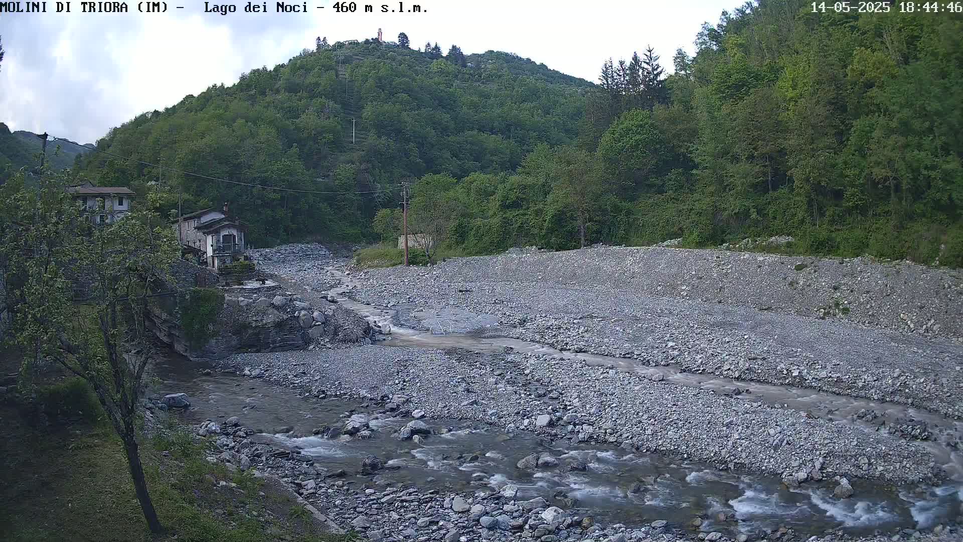 A rocky river flows through a valley, past two buildings nestled amongst lush green hills under a partly cloudy sky.