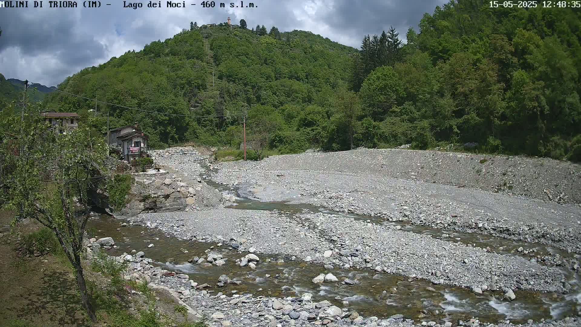A rocky stream flows through a valley under a partly cloudy sky, past a couple of buildings nestled beside a lush green hillside.