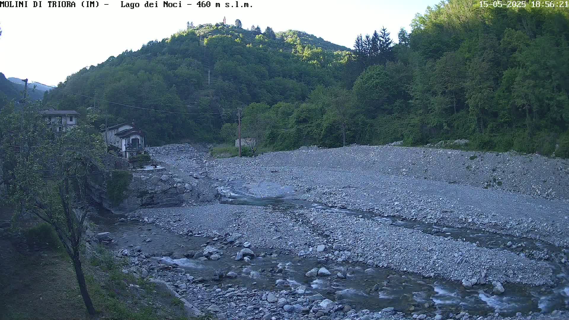 A rocky river flows through a valley under a twilight sky, past a couple of buildings nestled amongst green vegetation on a hillside.