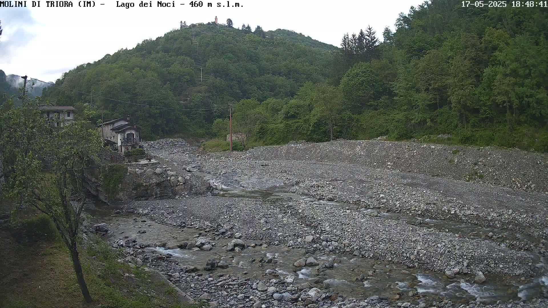 A rocky riverbed with a shallow stream flows through it, past buildings nestled at the base of a lush green hillside under an overcast sky.
