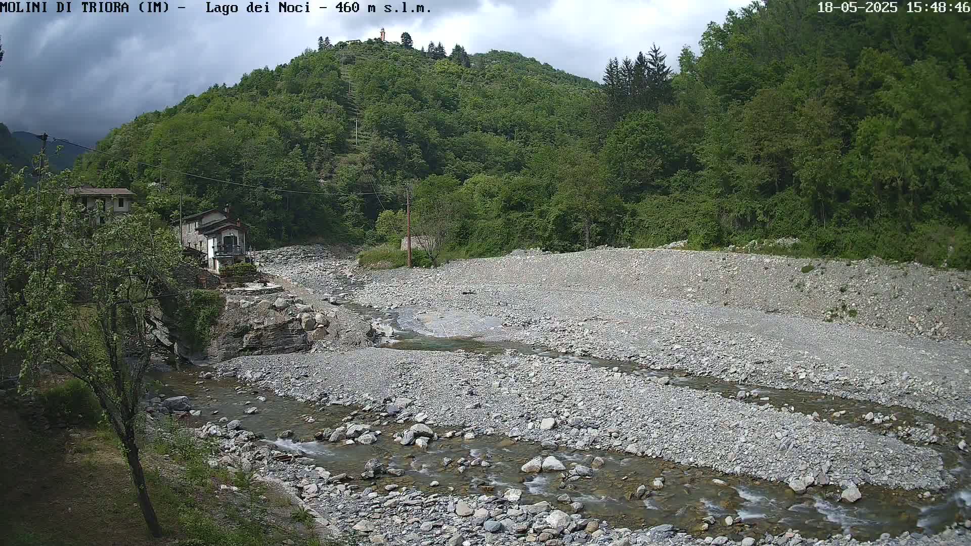 A rocky riverbed with a shallow, flowing stream cuts through a mostly dry riverbed, nestled beside a hillside covered in lush green trees under a partly cloudy sky.