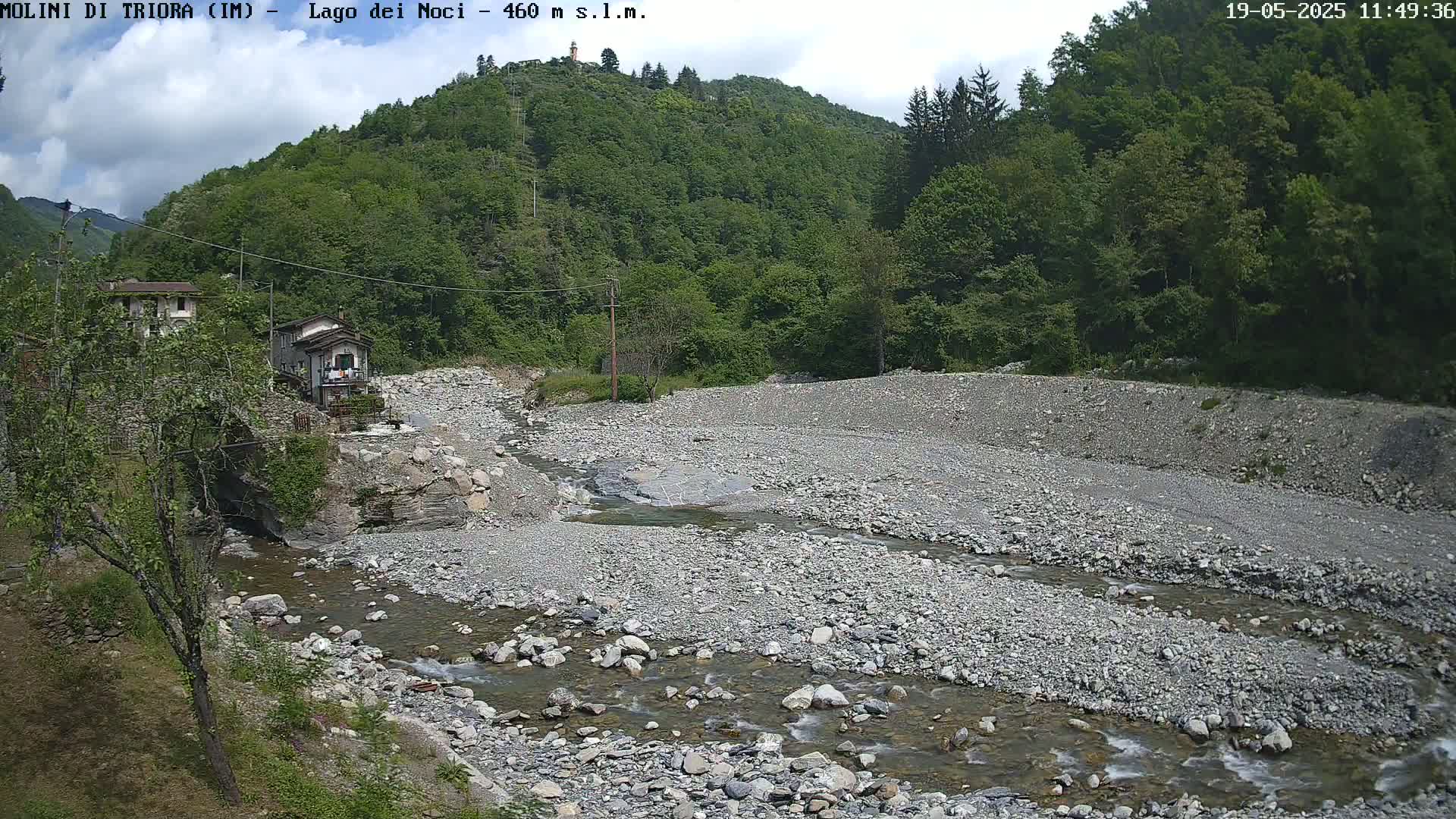 A rocky, shallow river flows through a dry riverbed, surrounded by lush green hills under a partly cloudy sky.