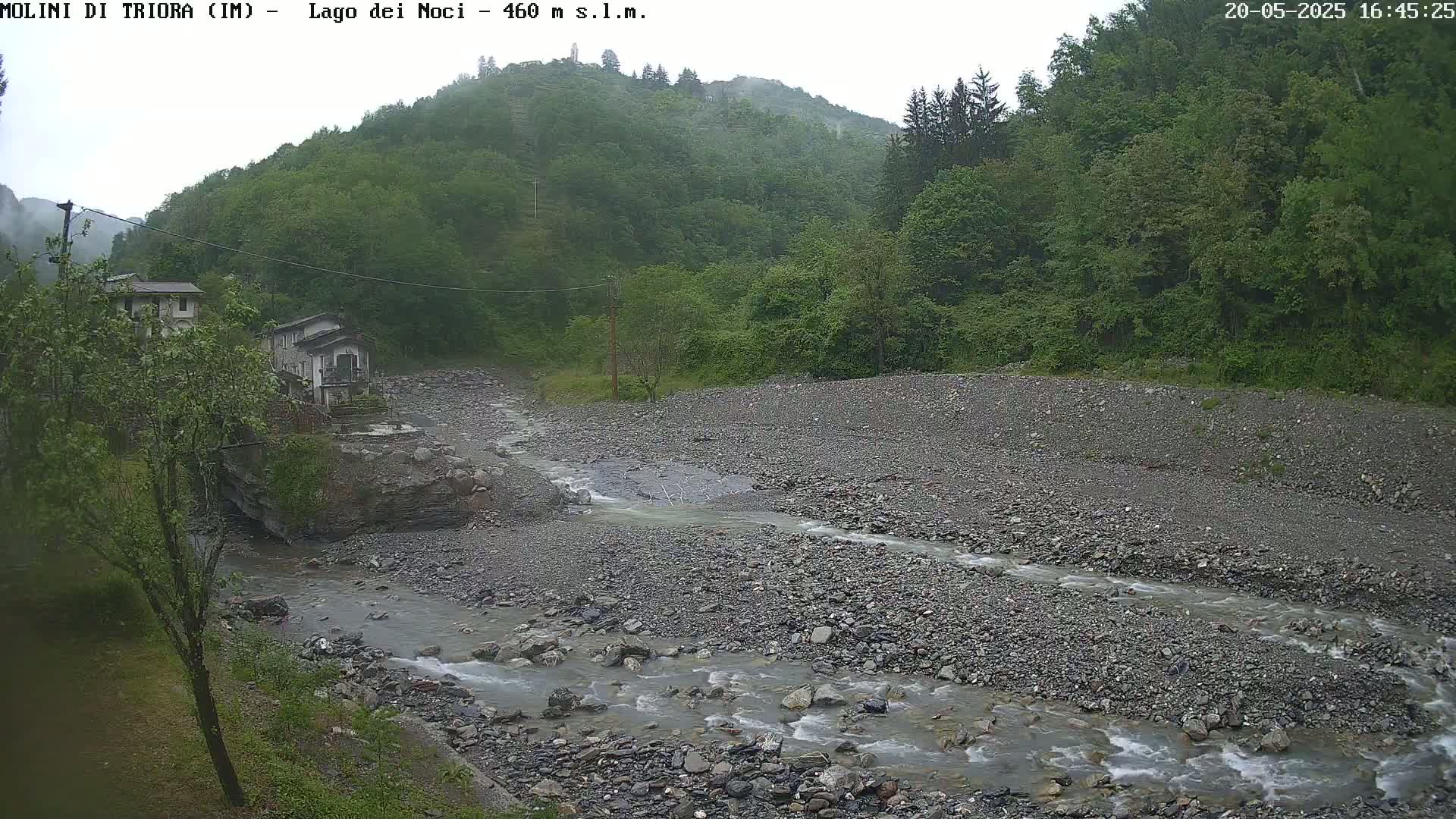 A rocky stream flows through a valley, past a couple of buildings and lush green hillsides, under an overcast sky.