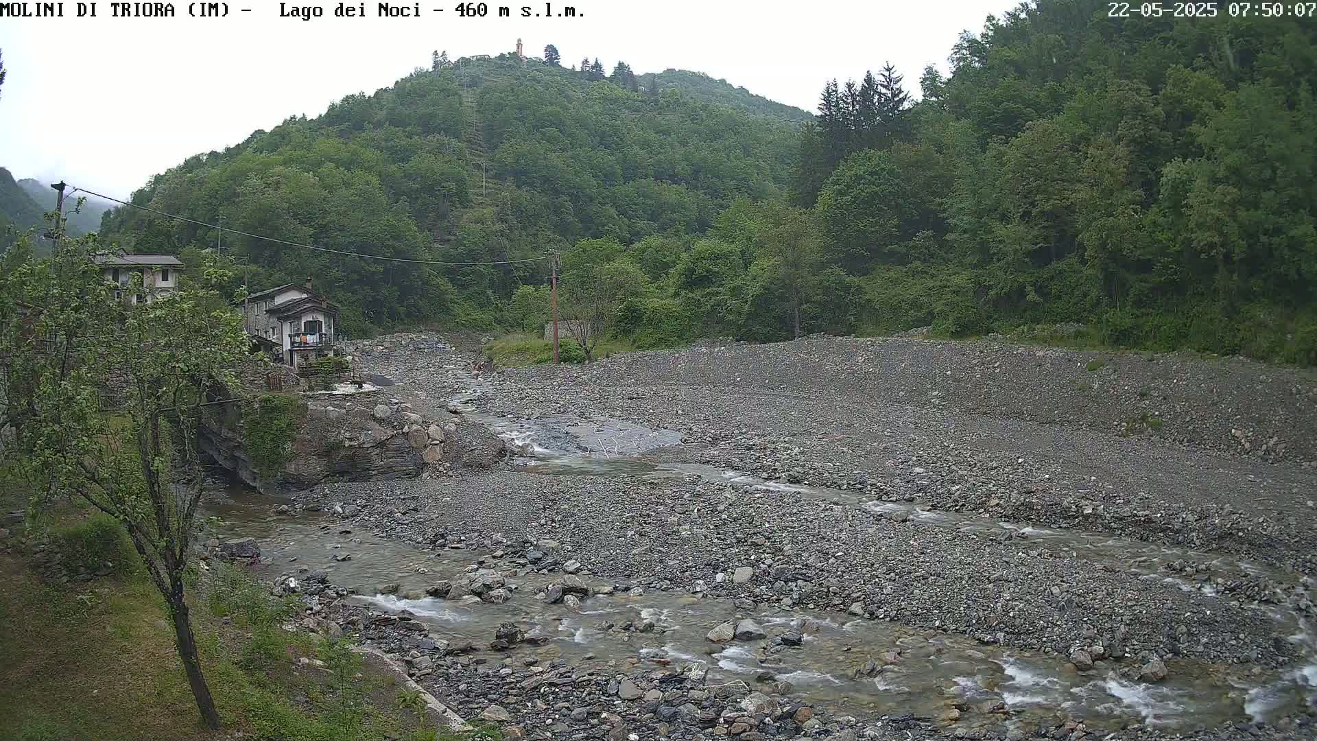 A rocky, shallow river flows through a valley on an overcast day, past a small village nestled beside a lush green hillside.