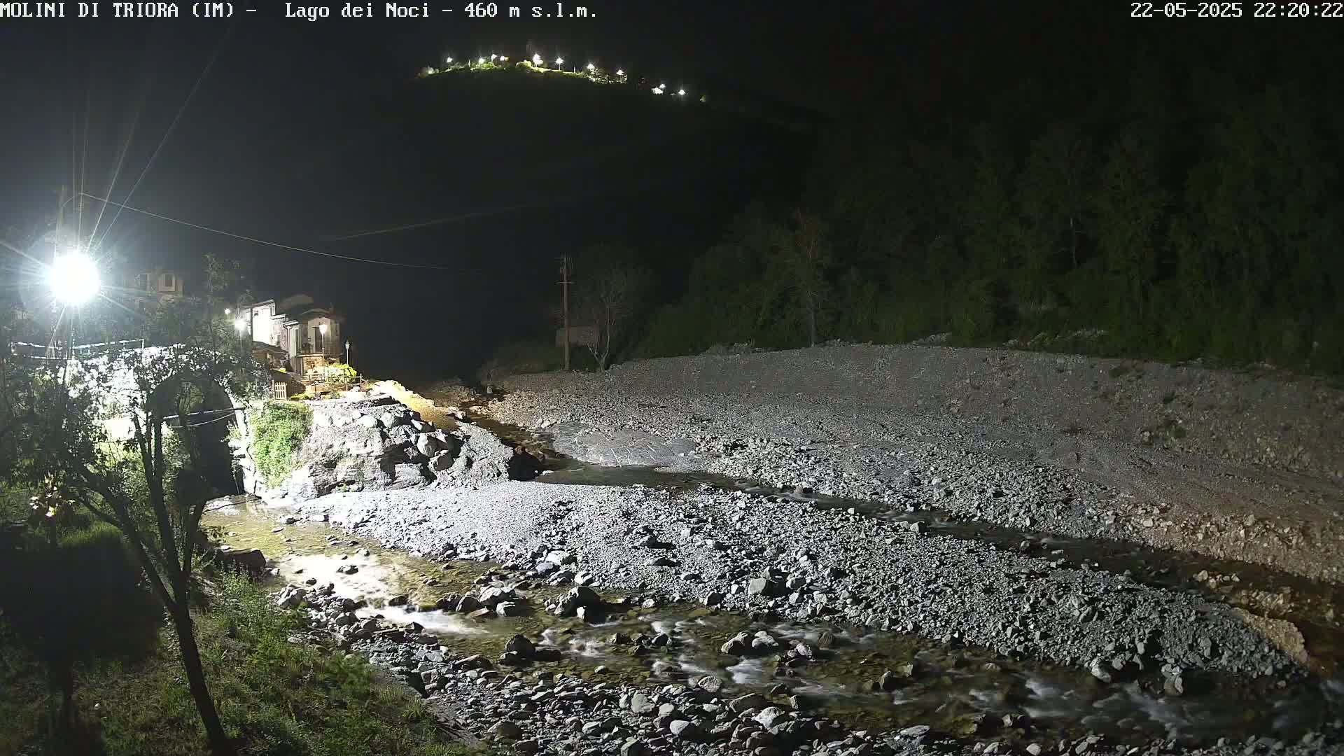 A rocky stream flows through a nighttime landscape, past a stone bridge and buildings under artificial light.