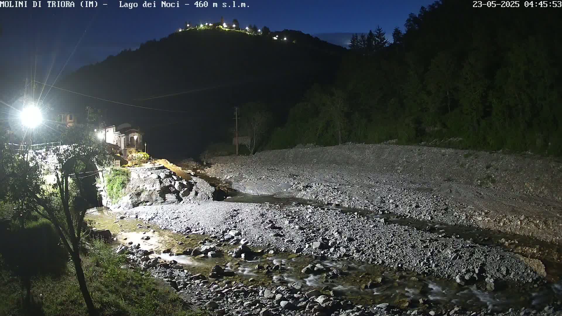A rocky stream flows through a dark, nighttime valley, past a stone bridge and a small village under a clear, moonlit sky.