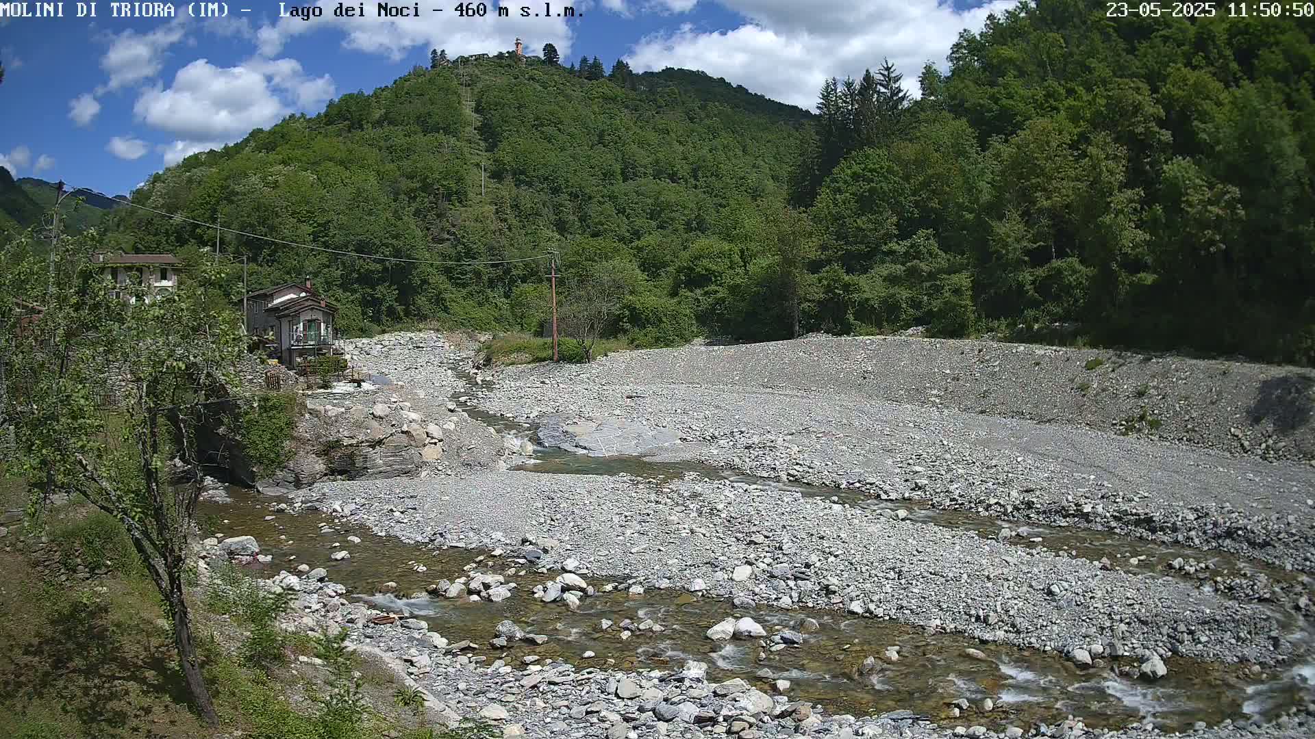 A rocky riverbed with a shallow, flowing stream winds through a valley under a partly cloudy sky, past a couple of buildings nestled among green, leafy trees on a hillside.