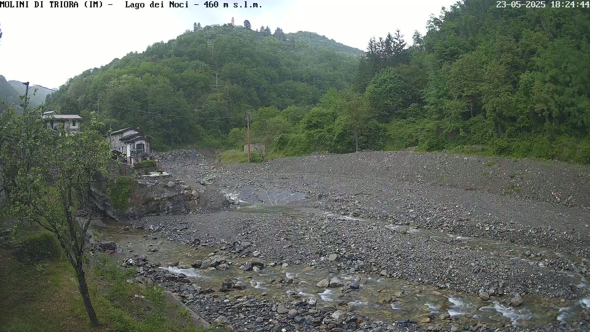 A rocky stream flows through a valley, past a few buildings nestled among lush green hills under an overcast sky.