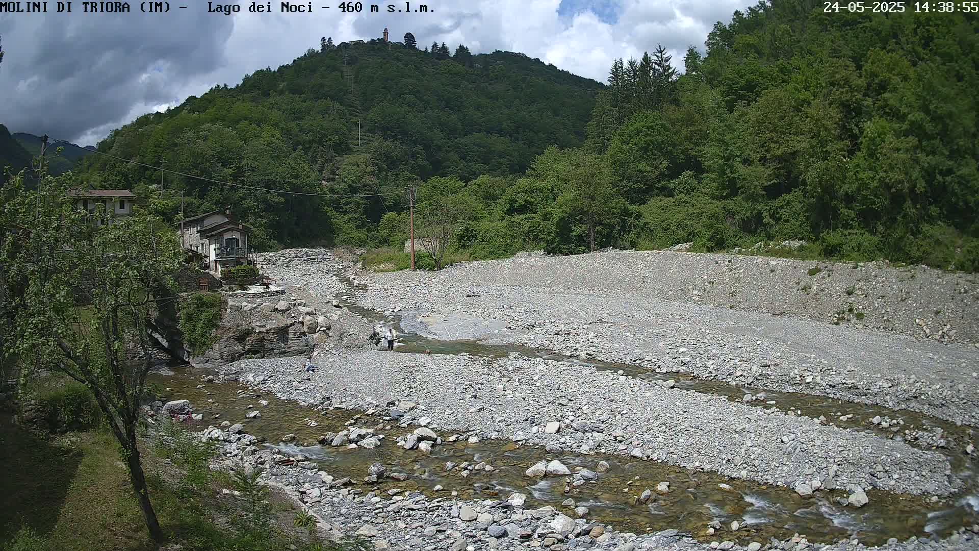 A partly cloudy day reveals a rocky, shallow riverbed with a slow-moving stream flowing through it, nestled between lush green hills and a small village.