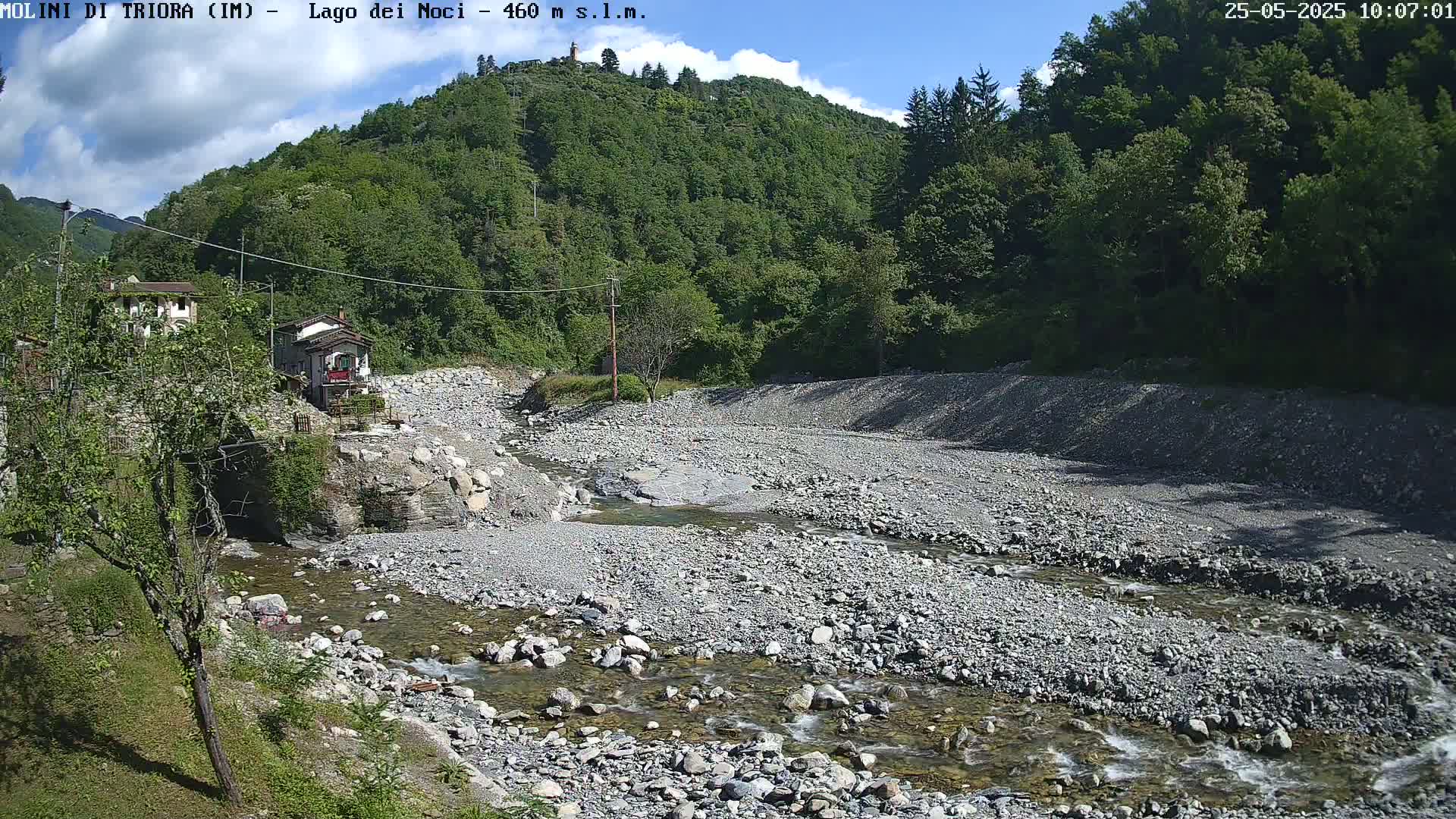 A rocky, shallow stream flows through a dry riverbed, surrounded by lush green hills under a partly cloudy sky.