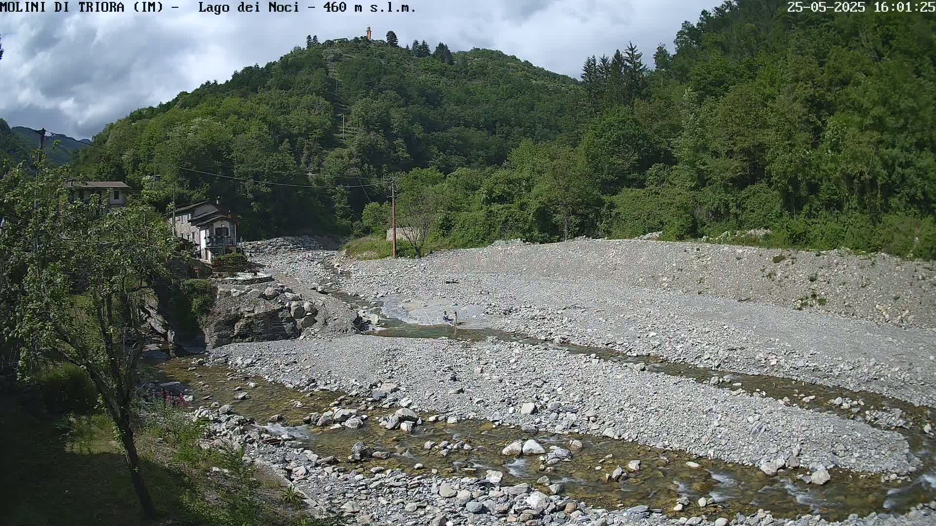 A rocky, shallow river flows through a dry riverbed, bordered by lush green vegetation and a small building, under a partly cloudy sky.