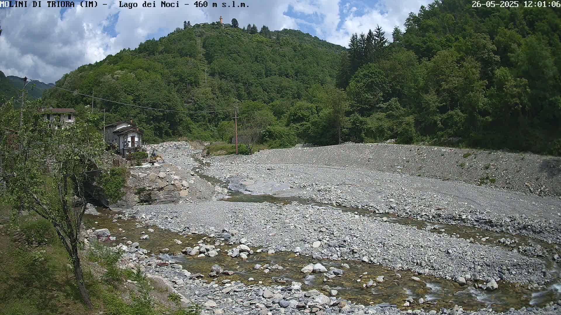 A rocky riverbed with a shallow stream flows through a valley under a partly cloudy sky, past buildings nestled at the base of a lush green mountain.