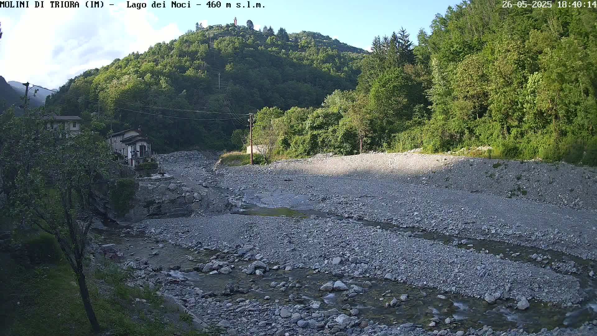 A rocky riverbed with a small stream flows through it, nestled in a valley surrounded by lush green vegetation under a mostly clear sky.