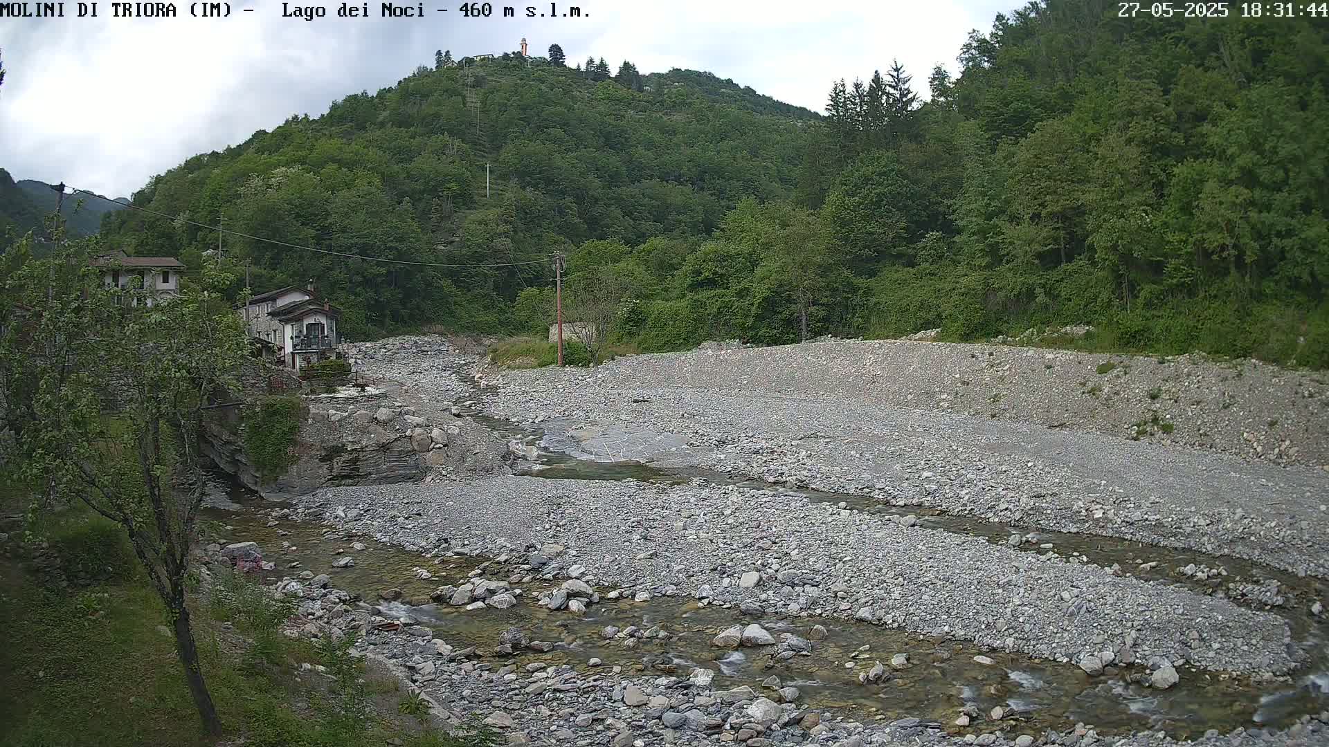 A rocky stream flows through a mostly dry riverbed, past buildings nestled at the base of a lush green hillside under a partly cloudy sky.