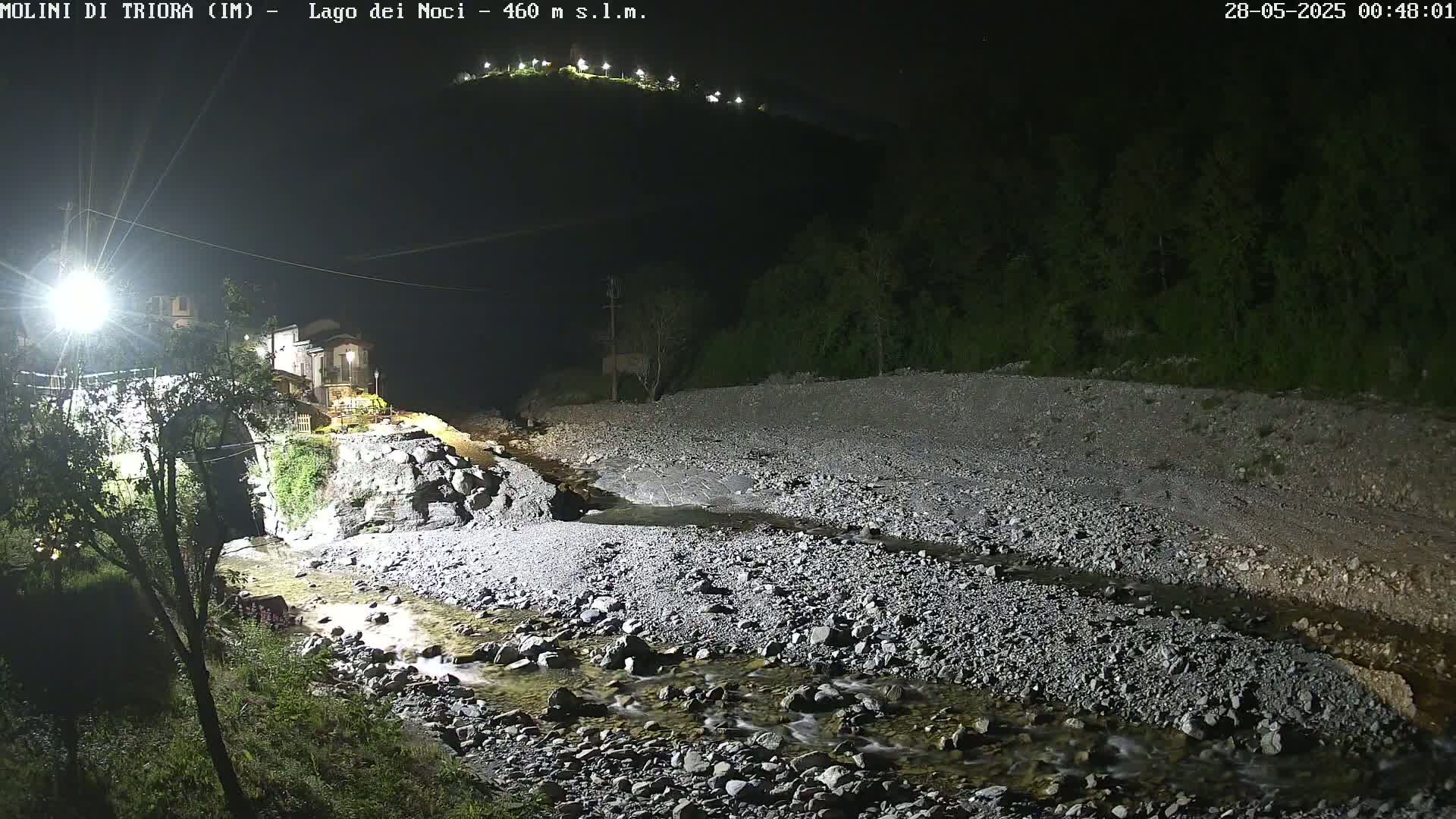 A rocky, shallow stream flows through a dark, nighttime landscape under artificial light, past a stone building and bridge near a hilltop with lights.