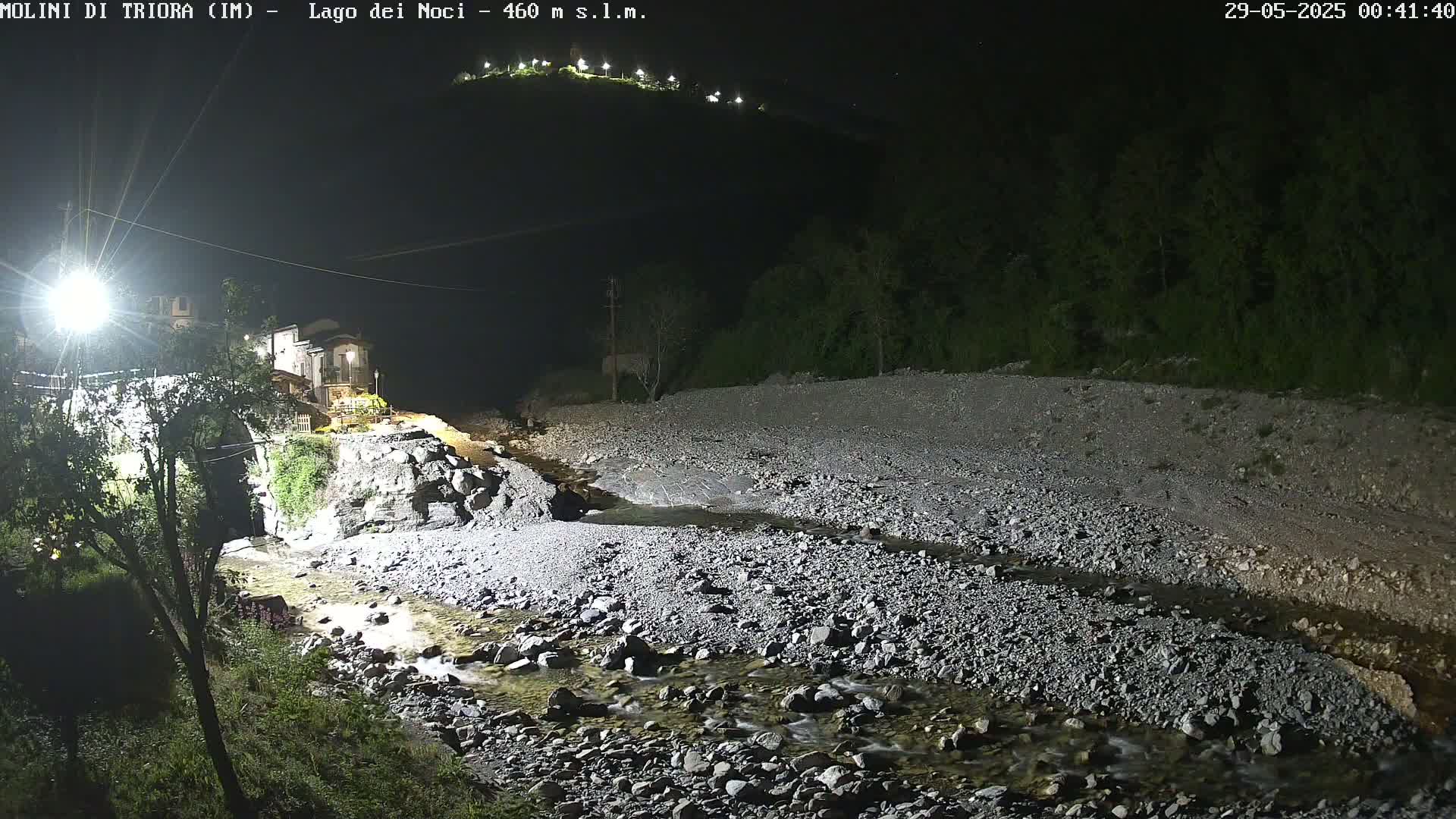 A rocky stream flows through a nighttime landscape, past a building and bridge, illuminated by artificial lights, under a clear, dark sky.