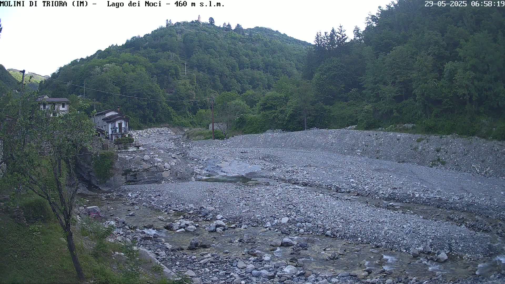 A shallow, rocky river flows through a valley, past a small village nestled against a lush, green hillside under an overcast sky.