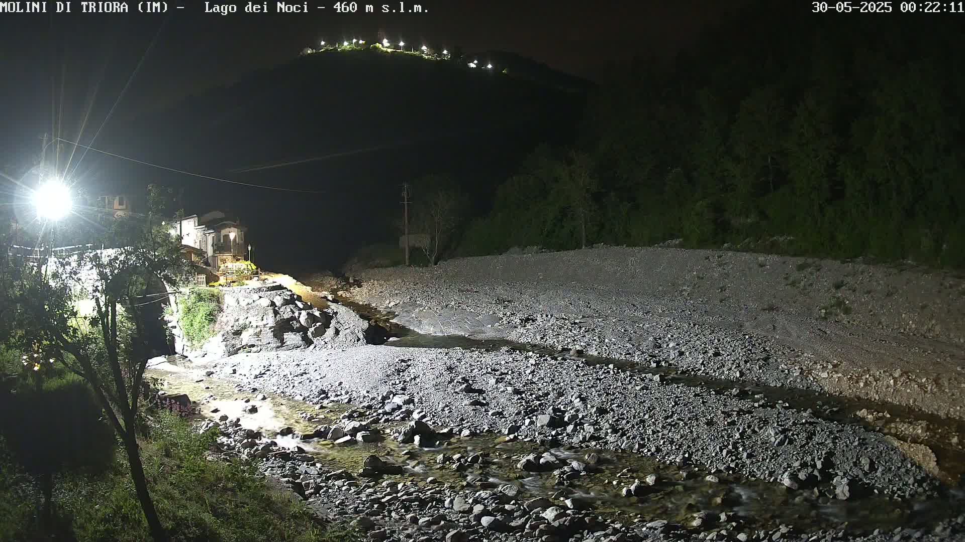A nighttime view of a rocky riverbed with a small stream flowing through it, near a village illuminated by artificial lights, under a clear night sky.