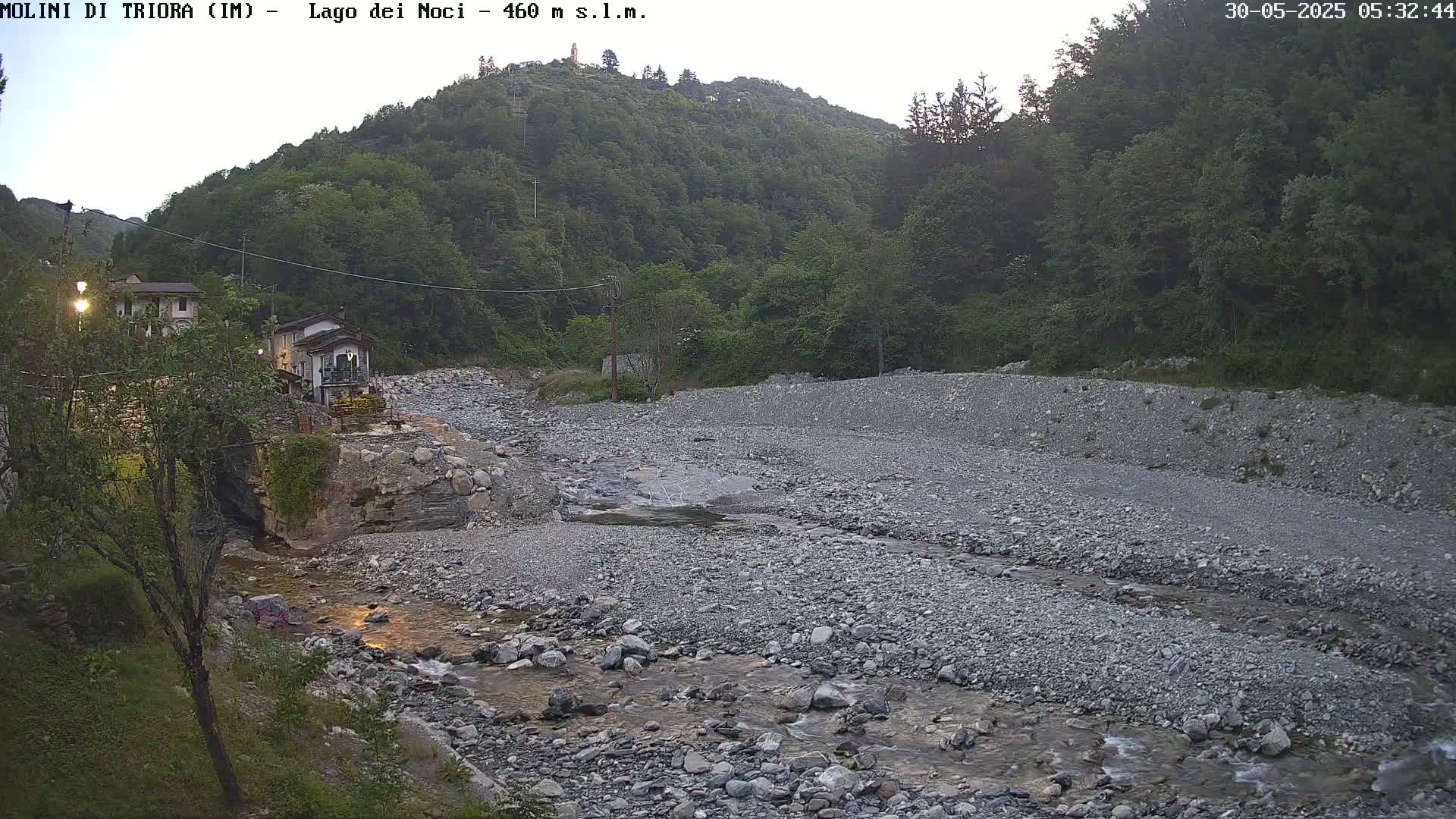 A rocky, shallow river flows through a valley under a dusky sky, past buildings nestled among lush green hills.