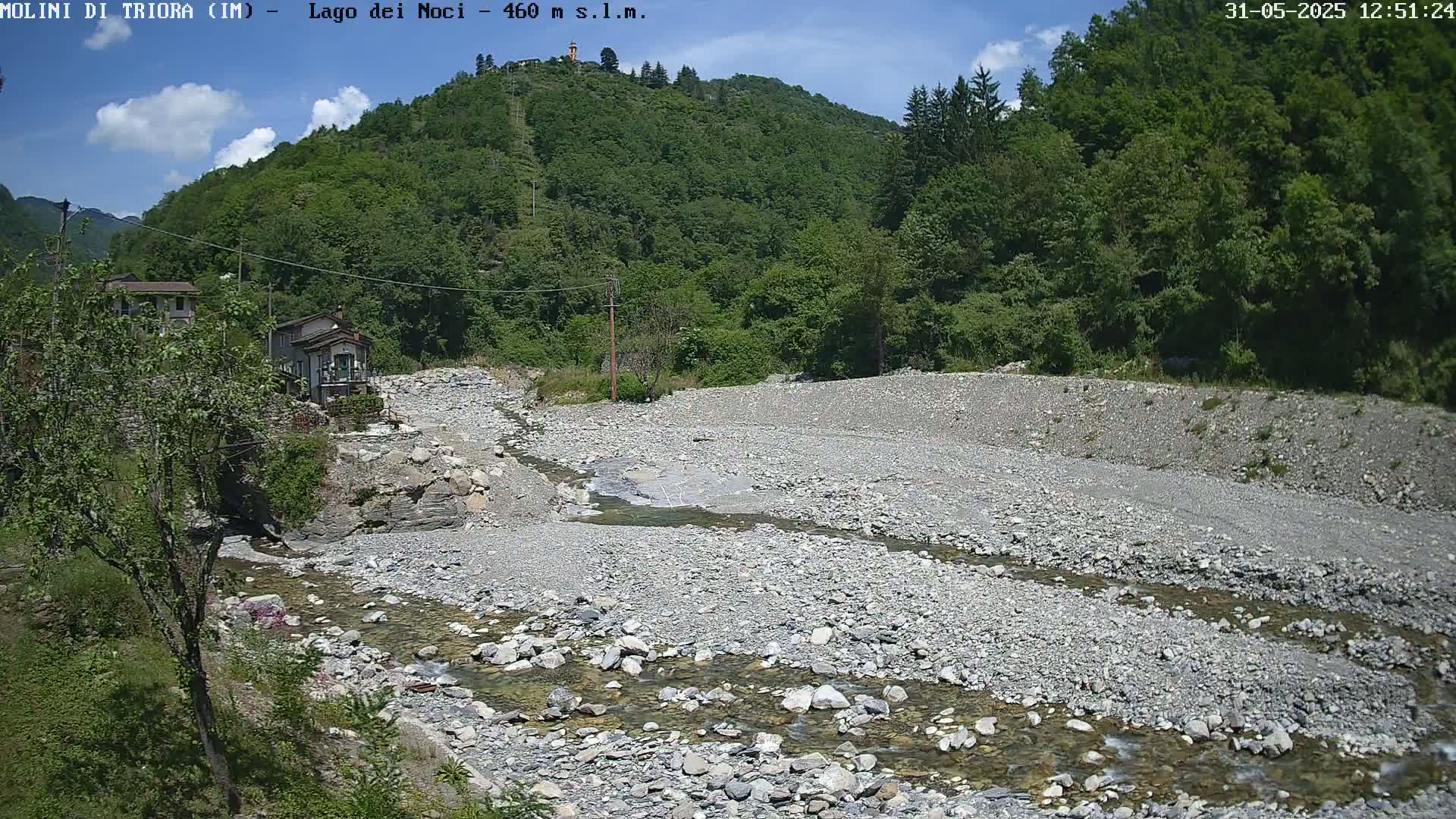 A shallow, rocky river flows through a dry riverbed, past some buildings nestled in a lush green hillside under a partly sunny sky.