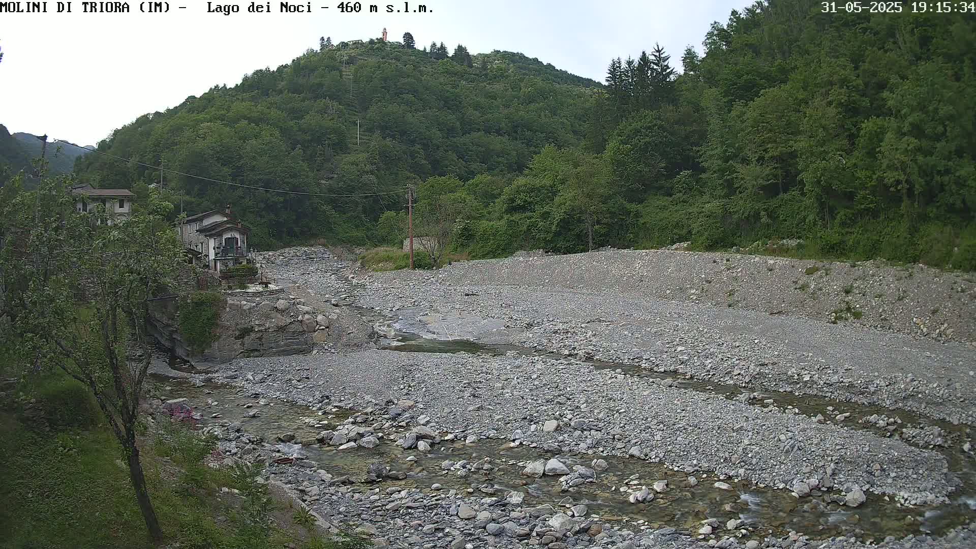 A rocky, shallow river flows through a valley, bordered by lush green vegetation and a few buildings under an overcast sky.