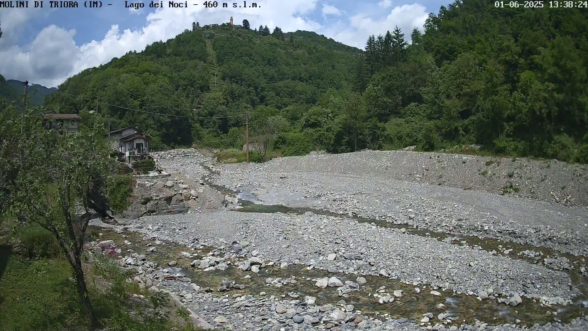 A shallow, rocky river flows through a dry, rocky riverbed, with lush green hills and a few buildings in the background under partly cloudy skies.